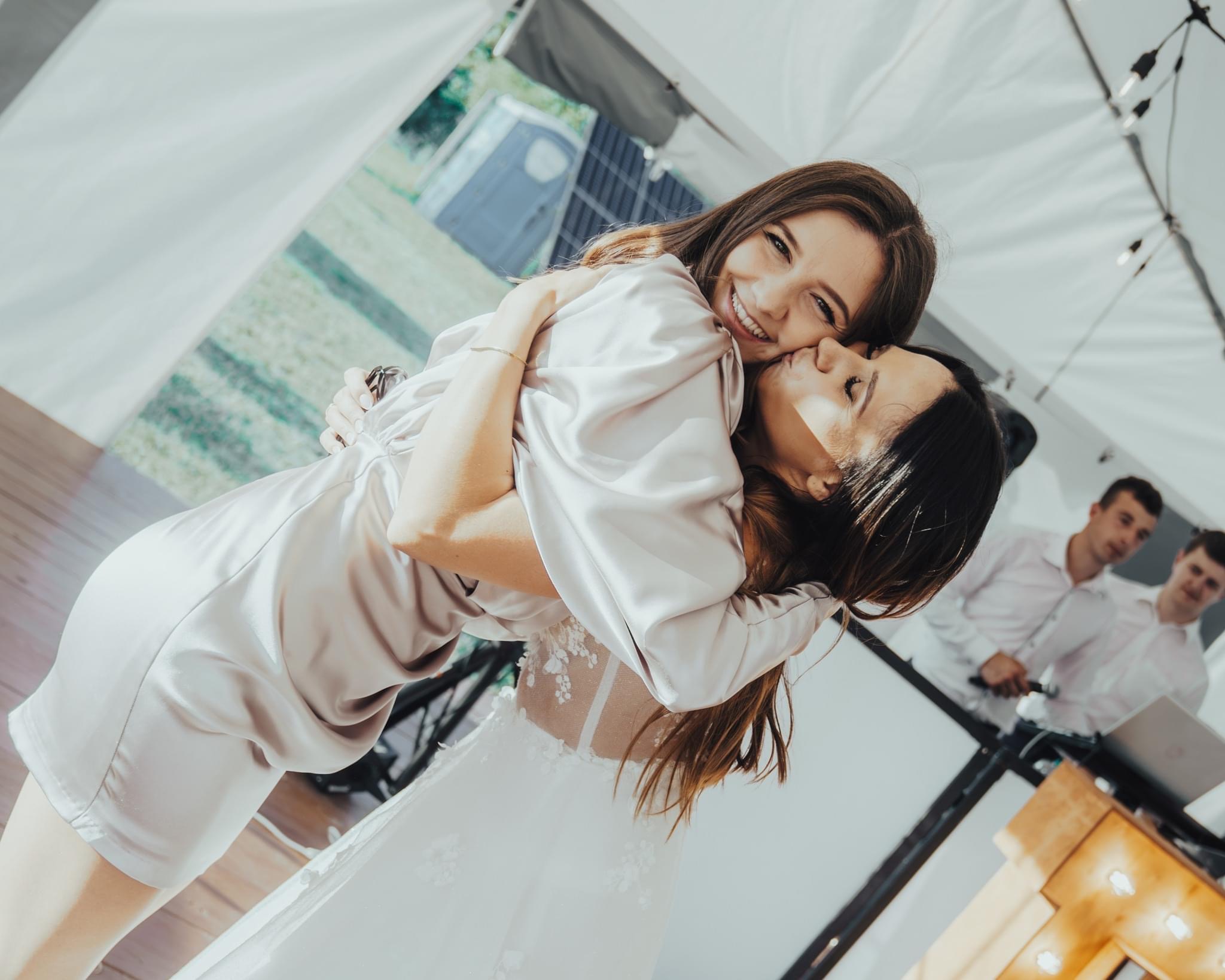 Panna młoda w białej sukni ślubnej przytula druhnę w satynowym szlafroku podczas wesela w namiocie, w tle widoczni dwaj mężczyźni.