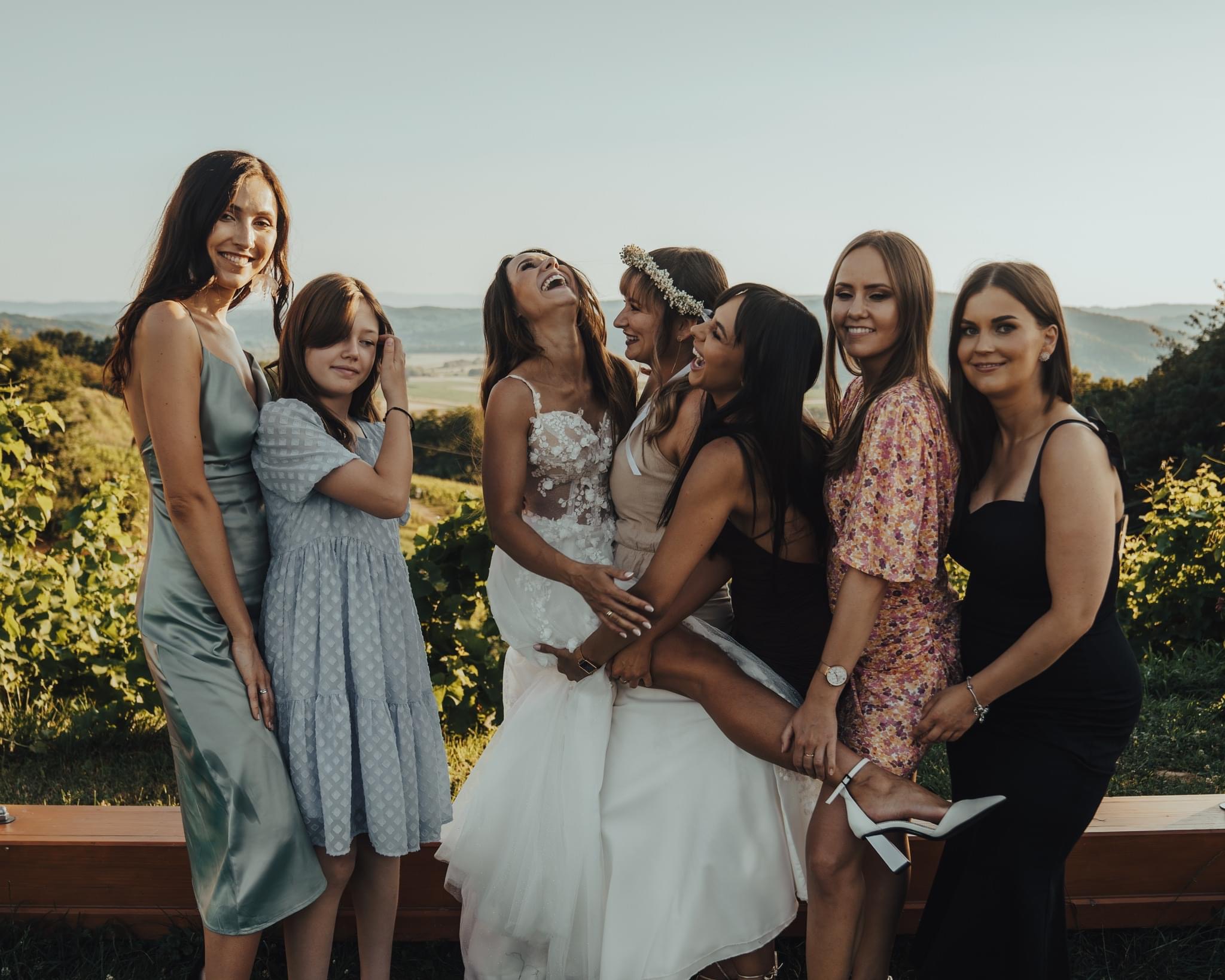 Panna młoda w białej sukni ślubnej, otoczona przez druhny w różnorodnych sukienkach, śmieje się z uniesioną głową, na tle zielonych wzgórz i jasnego nieba.
