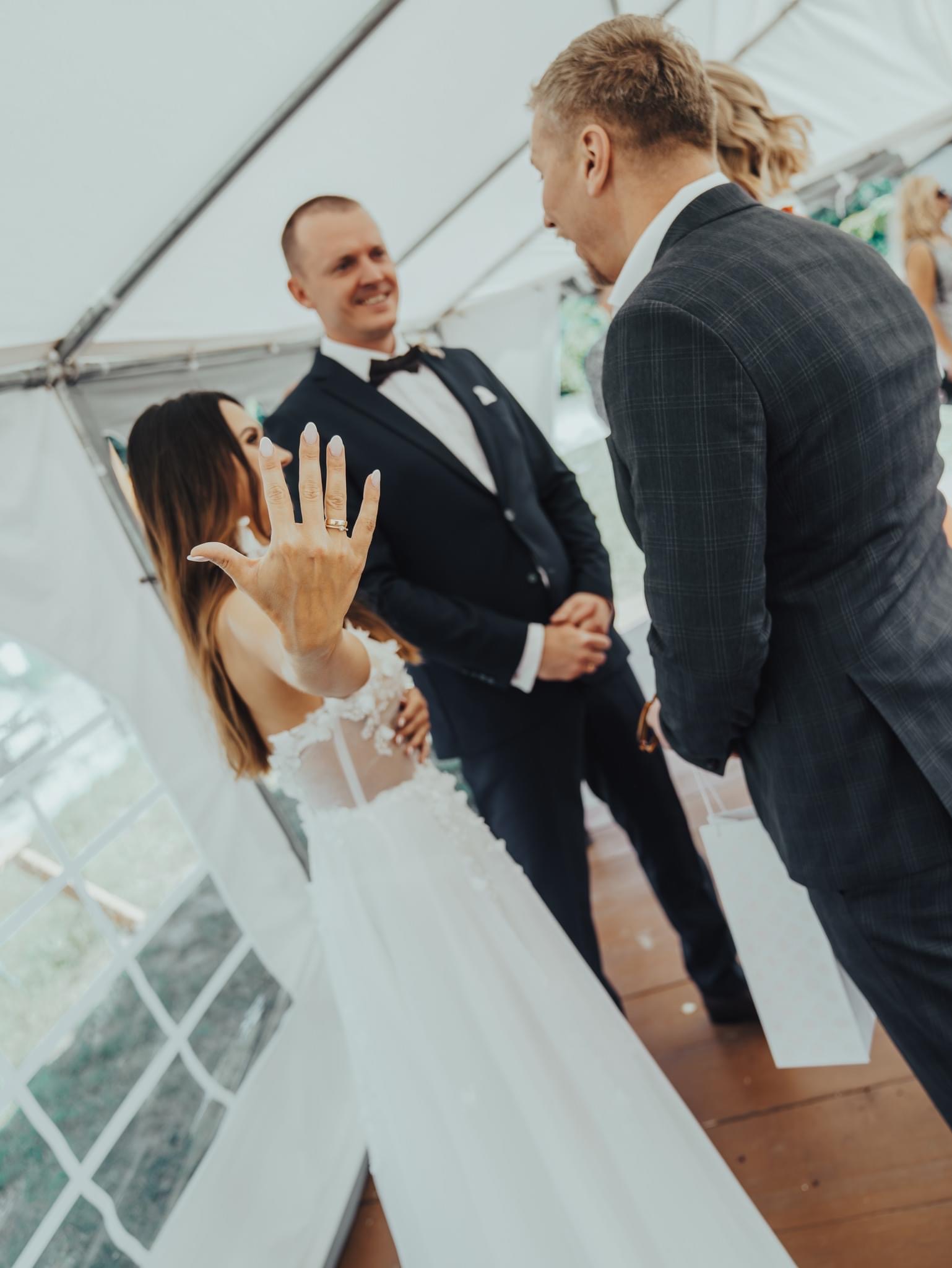 Panna młoda w białej sukni z koronkowymi aplikacjami, prezentuje pierścionek zaręczynowy na tle uśmiechniętego pana młodego w ciemnym garniturze i gościa w kraciastej marynarce.