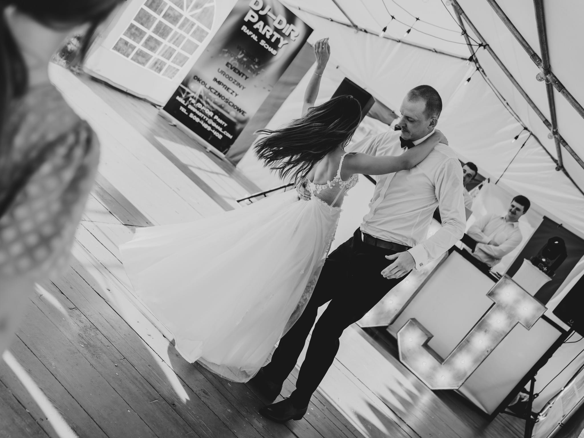 Czarno-białe zdjęcie pary młodej w tańcu na drewnianym parkiecie pod namiotem, z widocznym banerem DJ-a i oświetleniem dekoracyjnym.