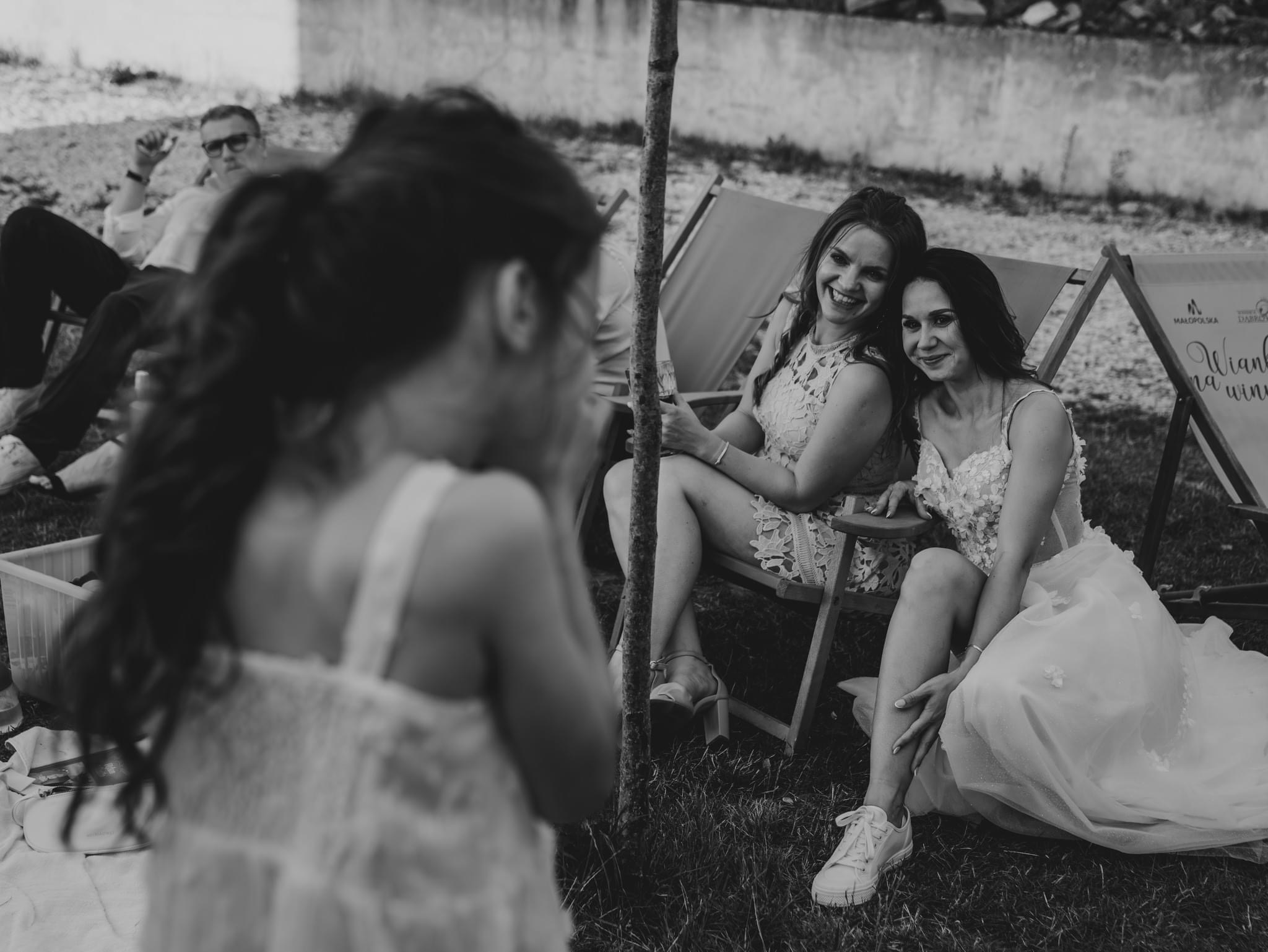 Dwie uśmiechnięte kobiety w sukienkach, jedna w koronkowej, druga w białej sukni ślubnej z kwiatowymi aplikacjami i białych tenisówkach, siedzą na leżakach na trawie, widok zza pleców dziecka,...