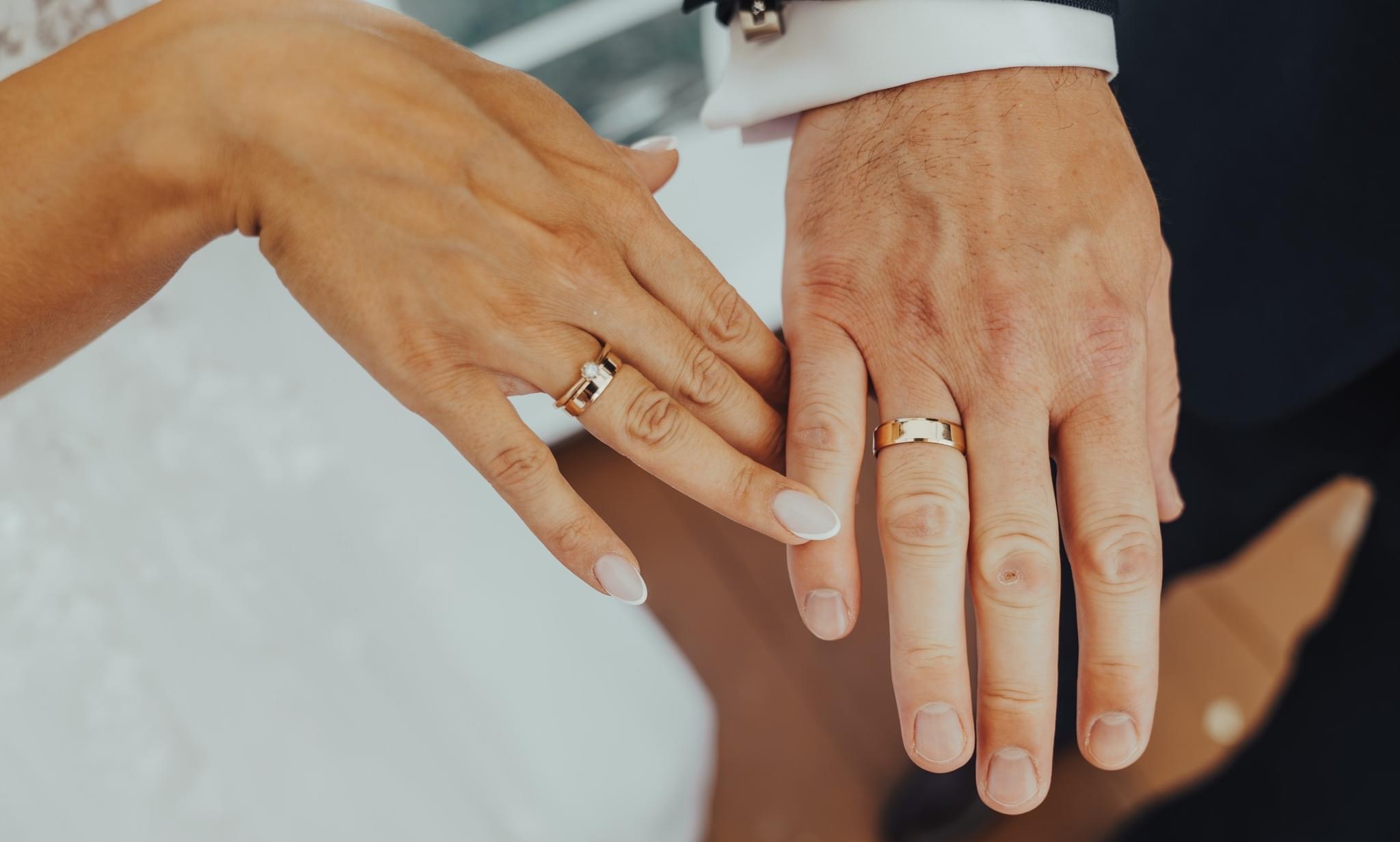 Dłonie nowożeńców z obrączkami: damska dłoń z pierścionkiem zaręczynowym i obrączką, męska dłoń z gładką, złotą obrączką, na tle białej sukni i fragmentu ciemnego garnituru.
