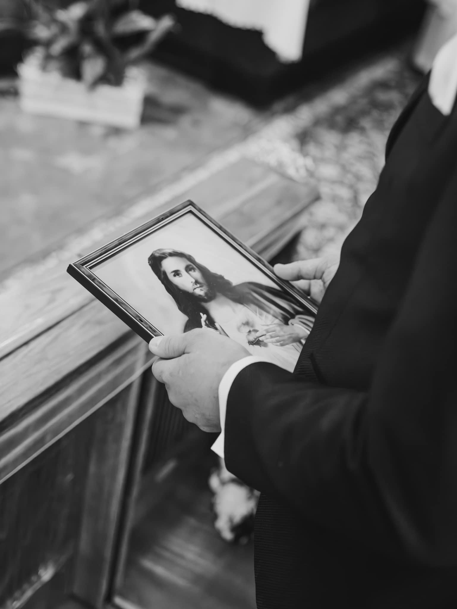 Czarno-białe zdjęcie mężczyzny w garniturze trzymającego oprawiony portret Jezusa Chrystusa w kościele.