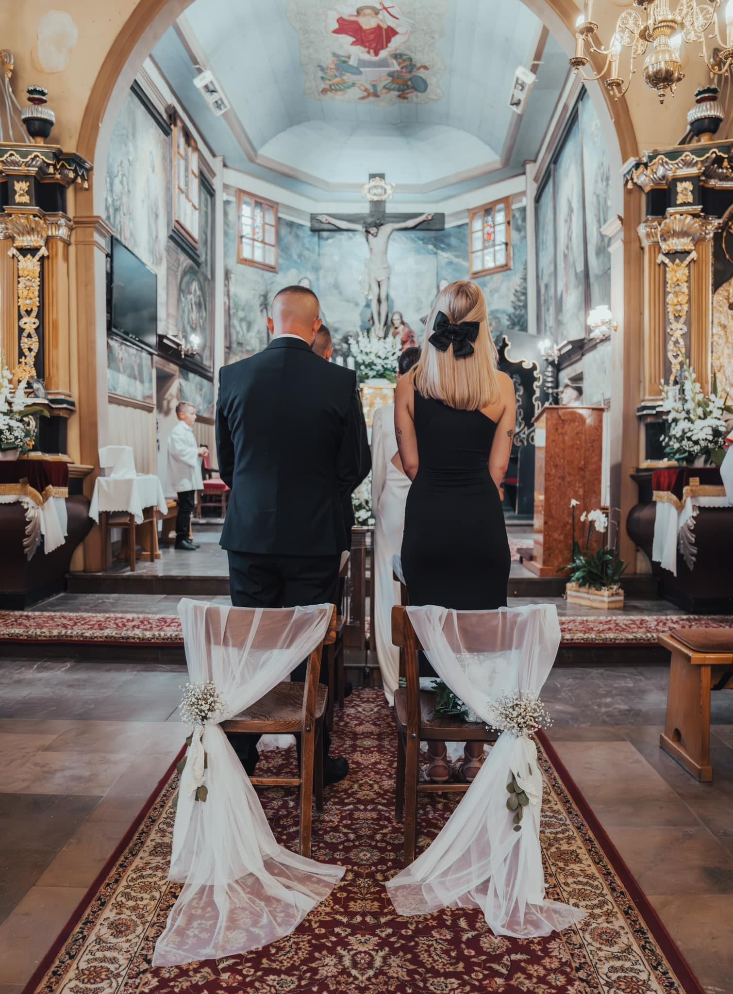 Para młoda podczas ceremonii w kościele z malowidłami na suficie, widok zza krzeseł udekorowanych białym tiulem i kwiatami.