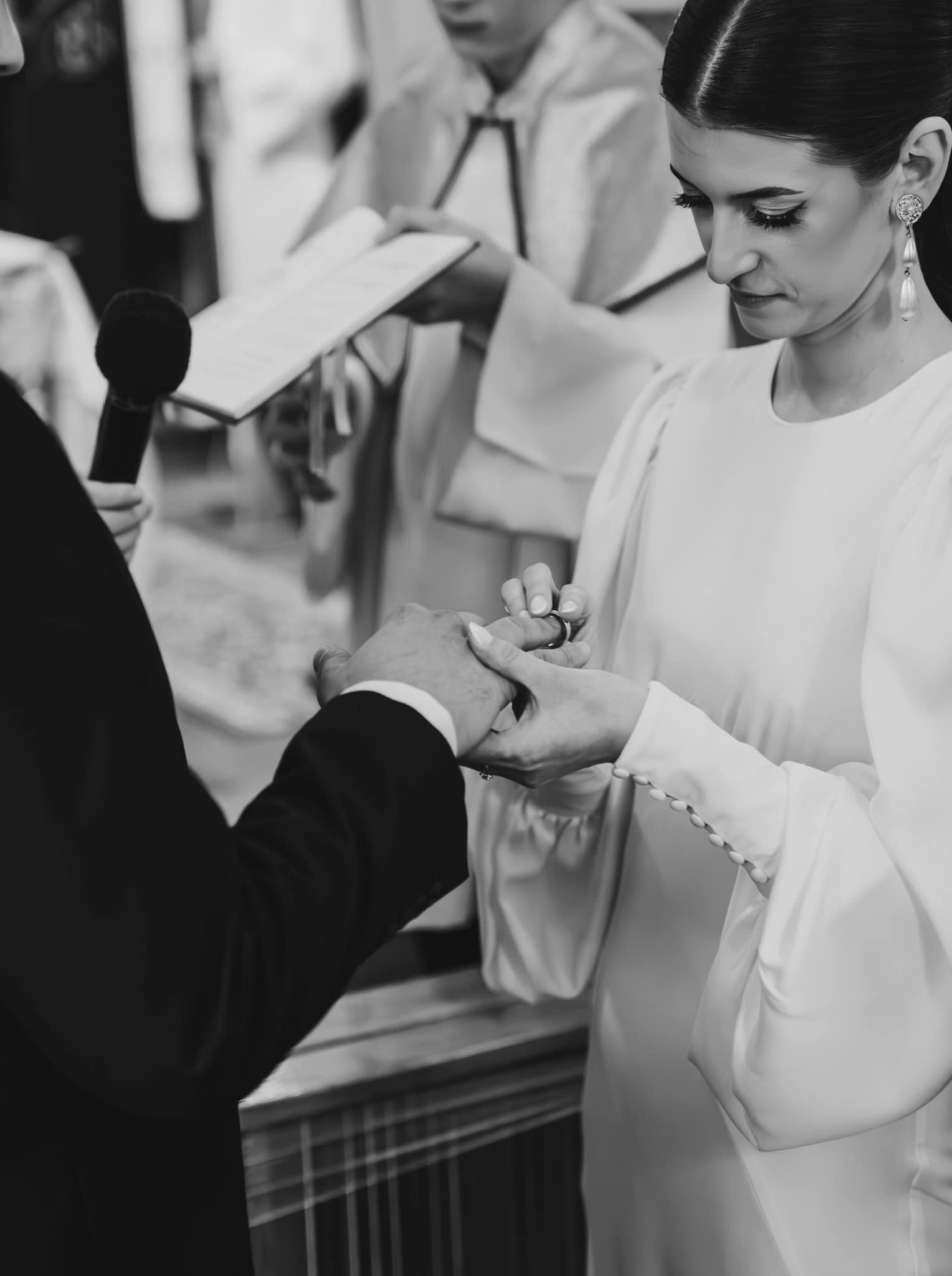 Czarno-białe ujęcie dłoni panny młodej wkładającej obrączkę na palec pana młodego podczas ceremonii ślubnej, z widocznym mikrofonem i osobą trzymającą księgę w tle.