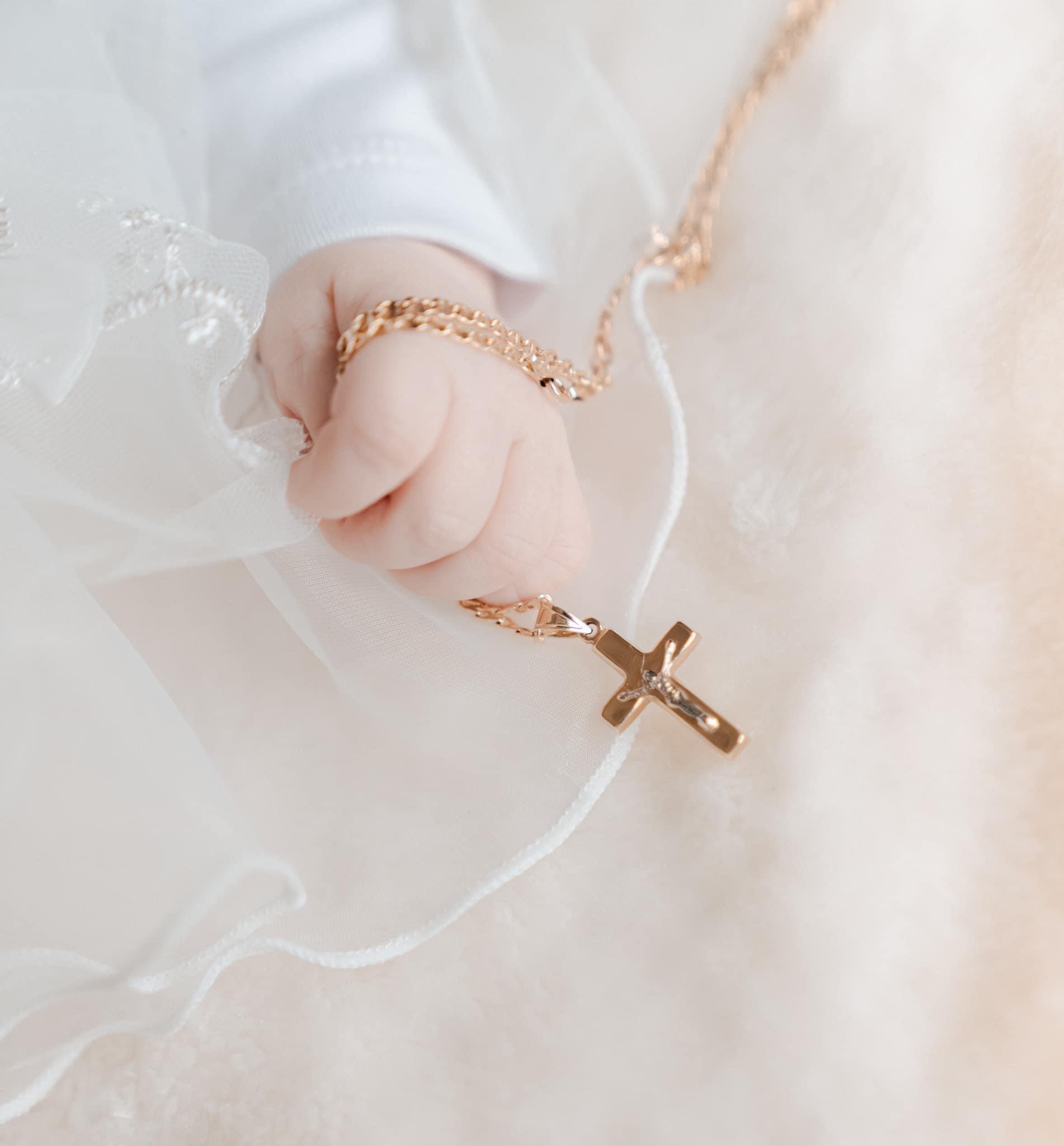 Dłoń niemowlęcia trzymająca złoty łańcuszek z krzyżykiem na tle białej, zwiewnej tkaniny.