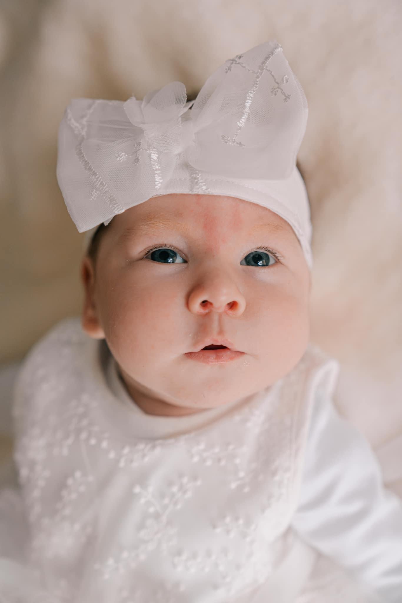 Portret niemowlęcia w białej sukience z koronkowym wzorem i opasce z tiulową kokardą, patrzącego w górę, z delikatnym oświetleniem.