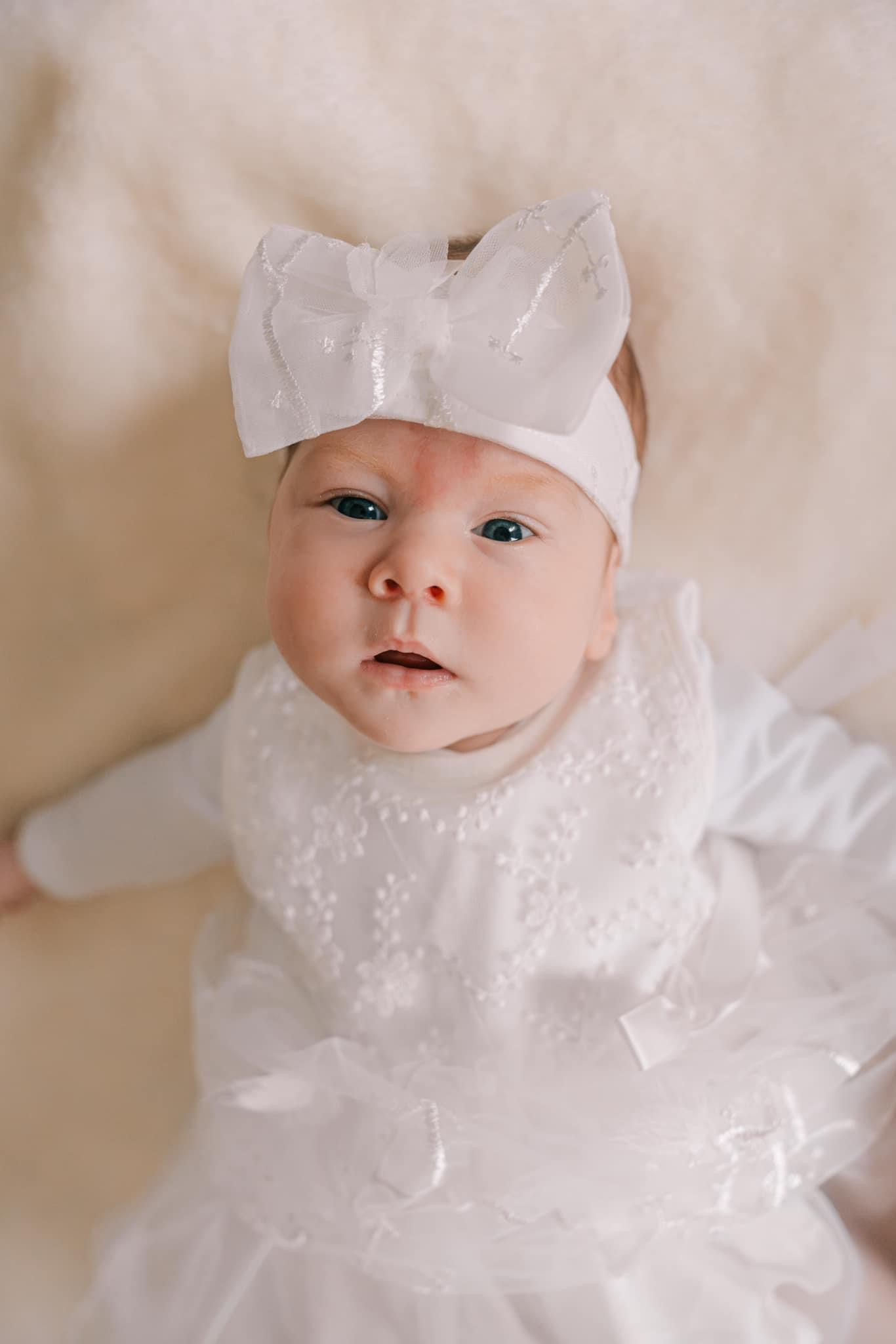 Niemowlę w białej sukience z koronkowymi zdobieniami i dużą kokardą na głowie, leżące na kremowym tle.