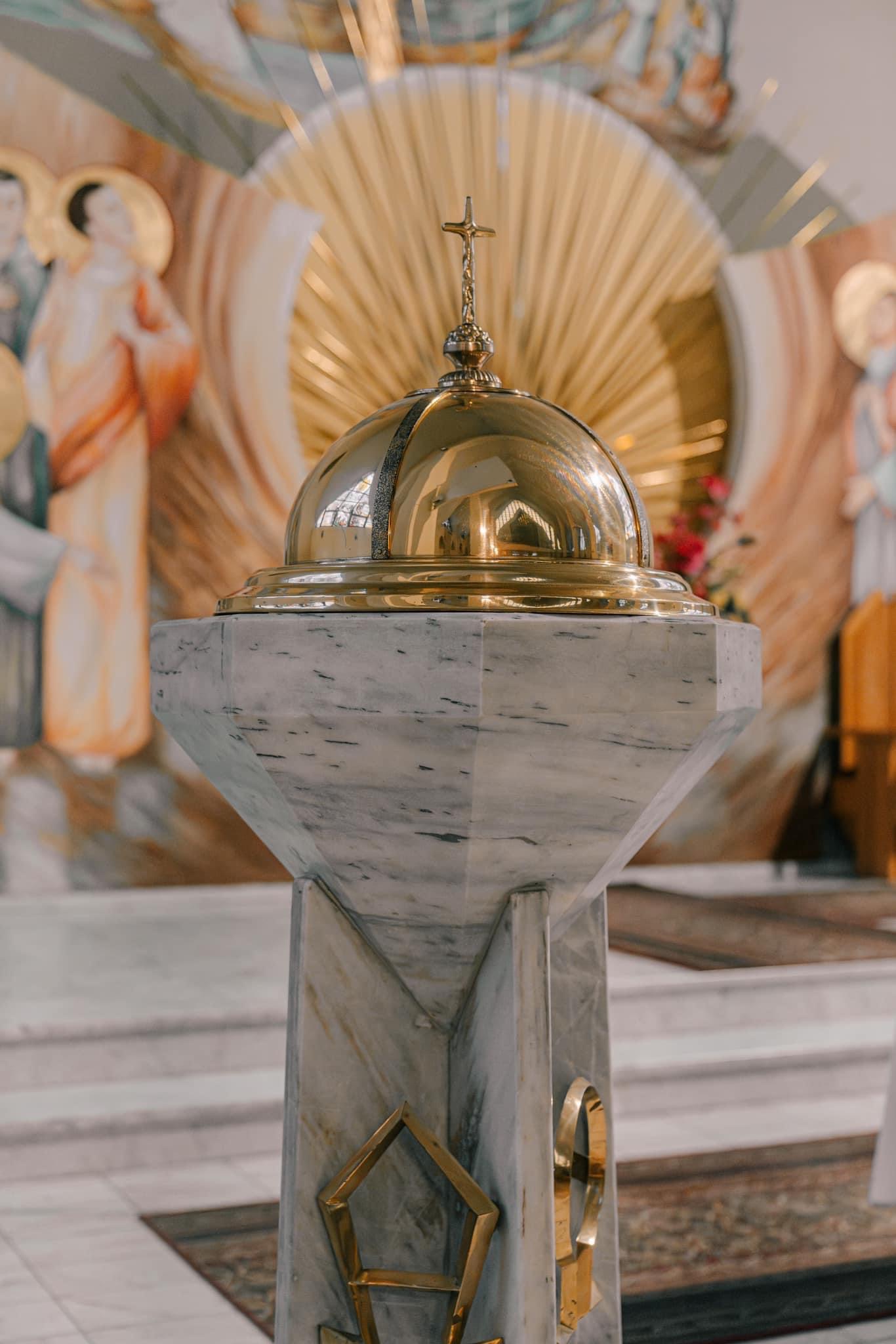 Marmurowa chrzcielnica z pozłacaną kopułą i krzyżem na tle fresków w kościele.