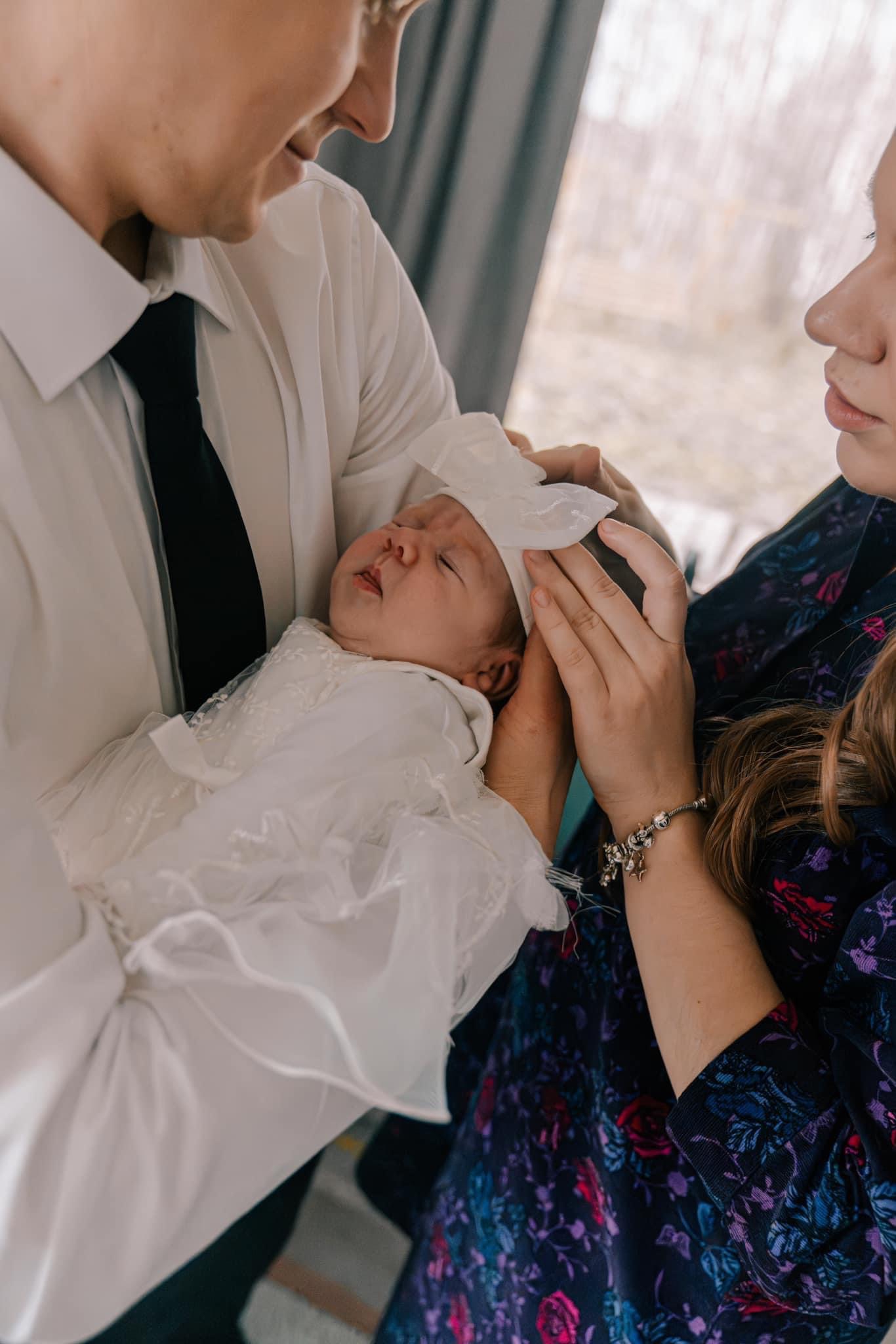 Rodzice trzymają i poprawiają czepek śpiącemu noworodkowi ubranemu w białą, koronkową sukienkę.