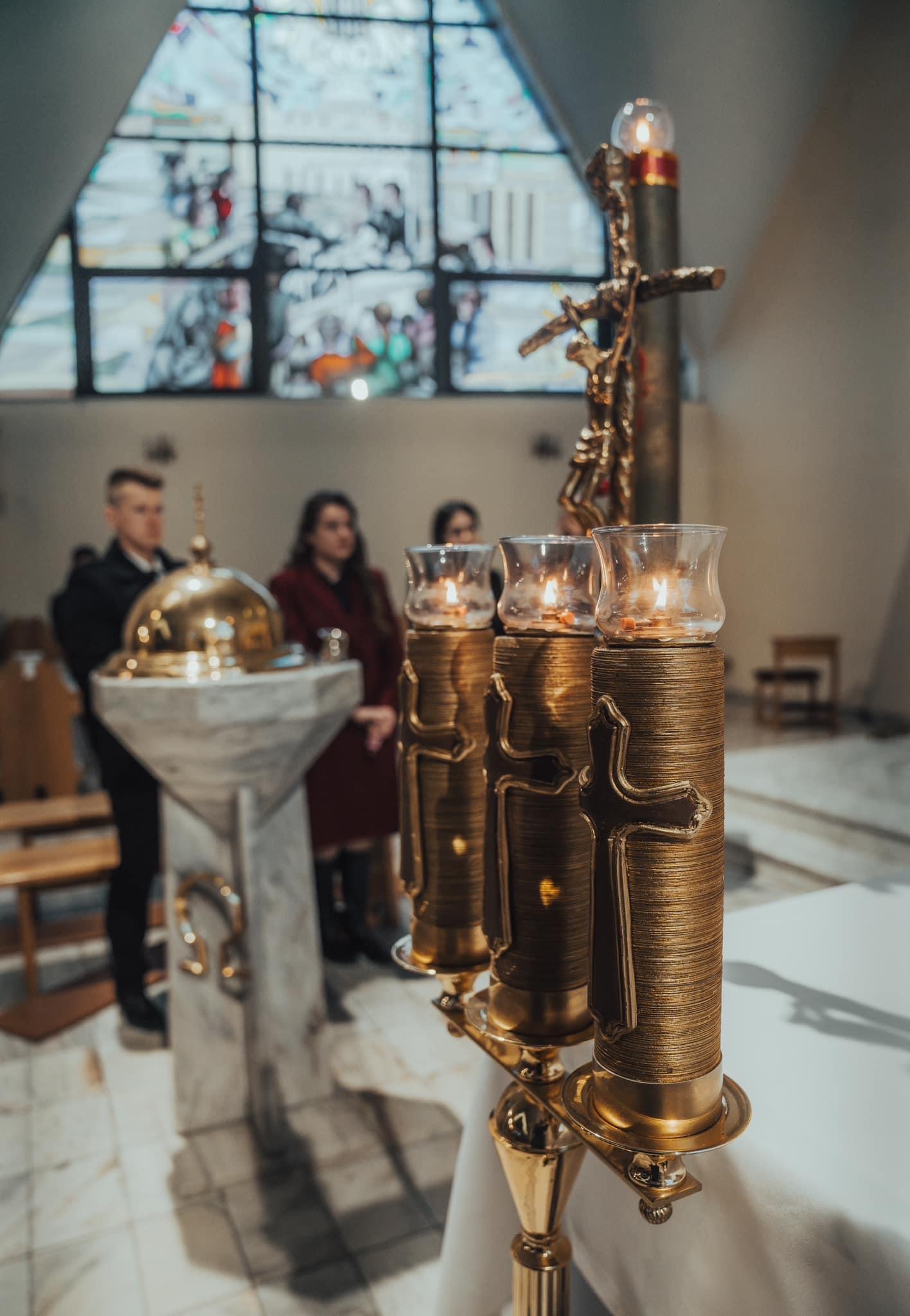Złote świeczniki z płonącymi świecami na białym obrusie, w tle osoby i witrażowe okno w kościele.