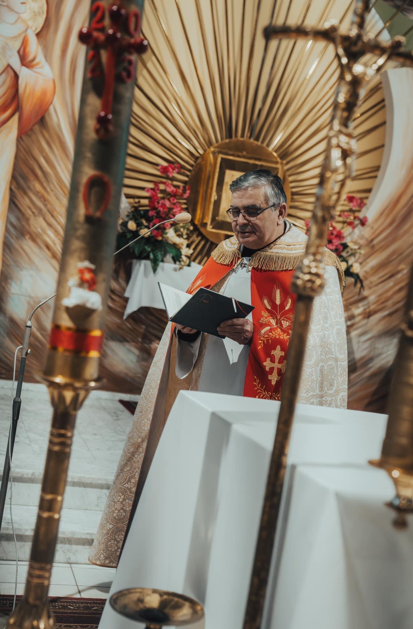 Ksiądz w ornacie, czytający z księgi podczas ceremonii w kościele, z widocznym krzyżem i świecami.