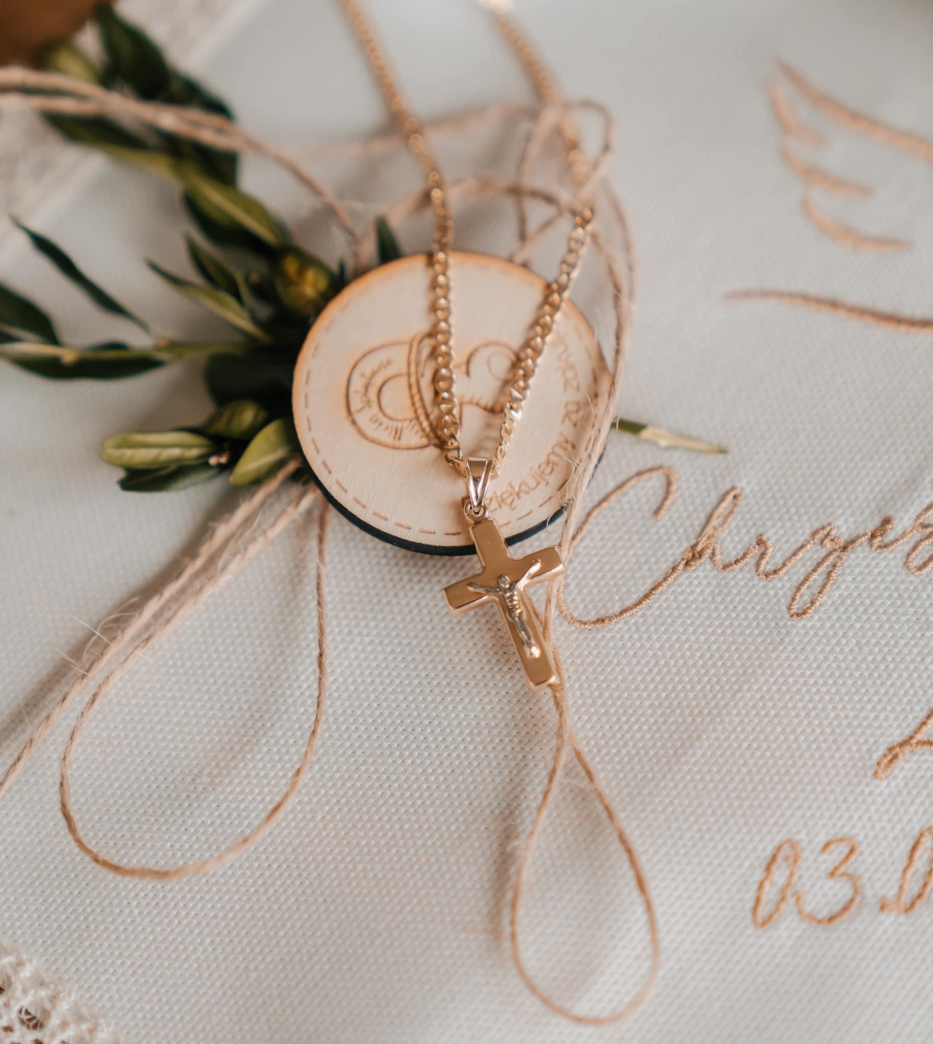 Złoty łańcuszek z krzyżykiem leżący na białej poduszce z haftowanym napisem i datą, obok drewnianego krążka z wygrawerowanymi obrączkami i gałązką oliwną, całość owinięta jutowym sznurkiem.