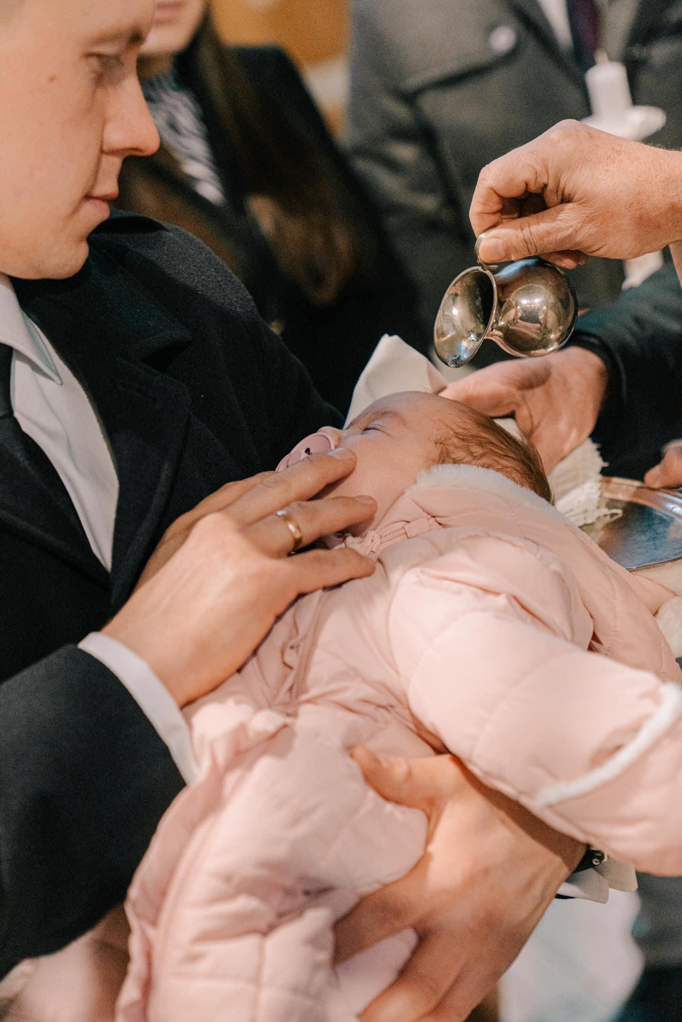 Niemowlę w różowym kombinezonie podczas chrztu, w tle widoczna srebrna konewka i dłoń z obrączką.