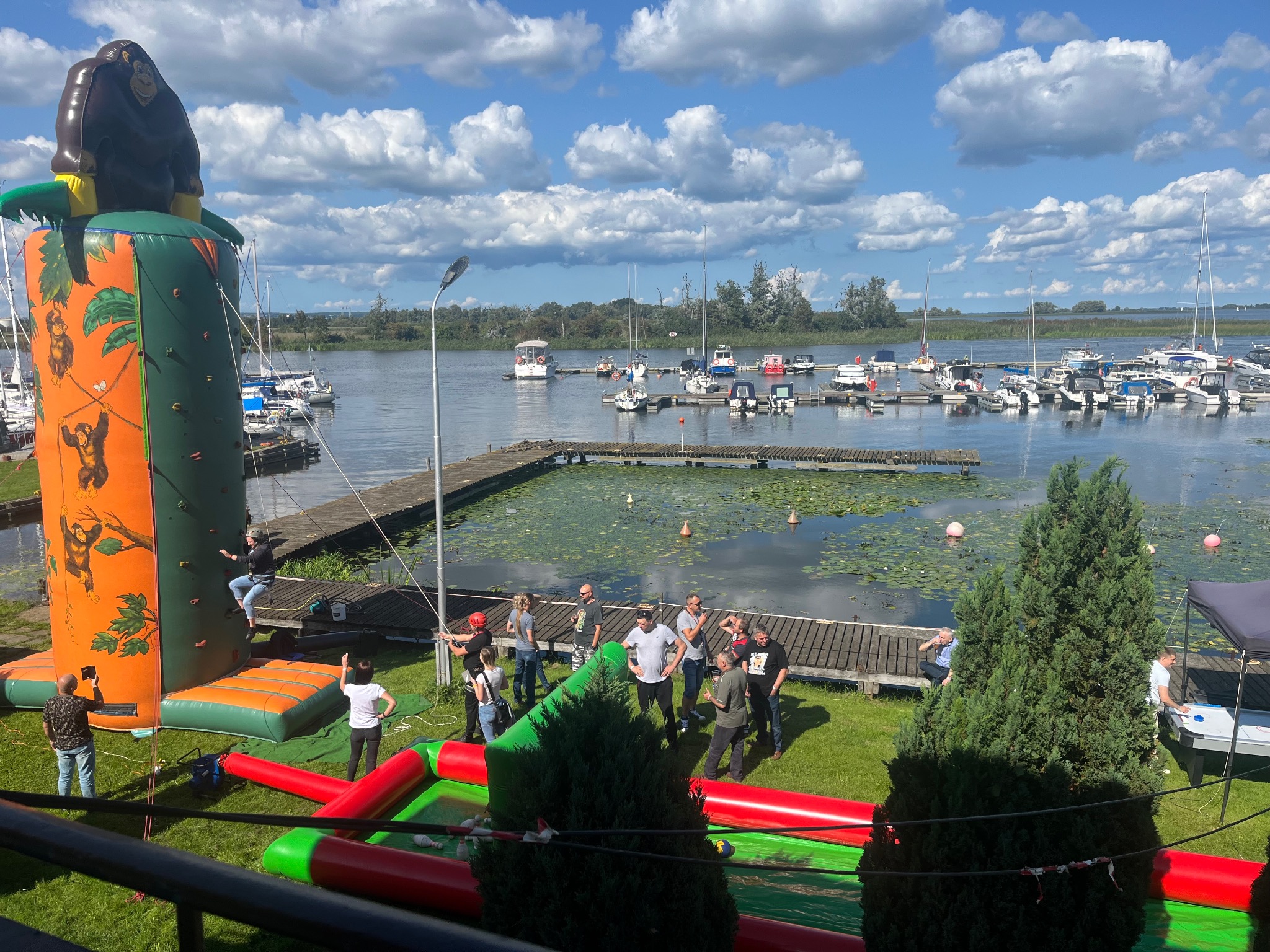 Grupa osób uczestniczy w imprezie plenerowej z dmuchaną ścianką wspinaczkową z motywem goryla, na tle jeziora i przystani jachtowej.