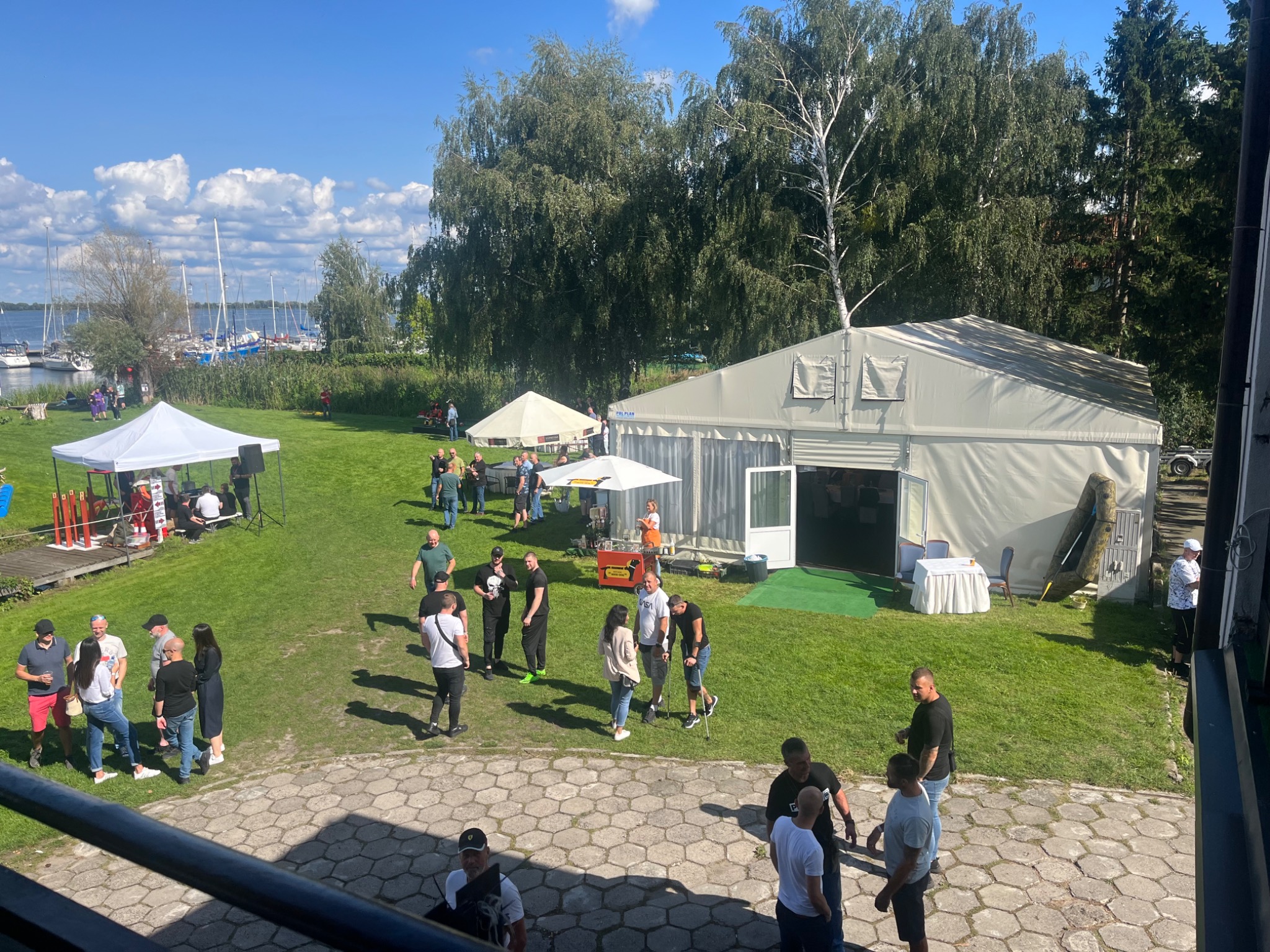 Plenerowa impreza z białymi namiotami i widokiem na jezioro w słoneczny dzień, uczestnicy w swobodnych strojach.