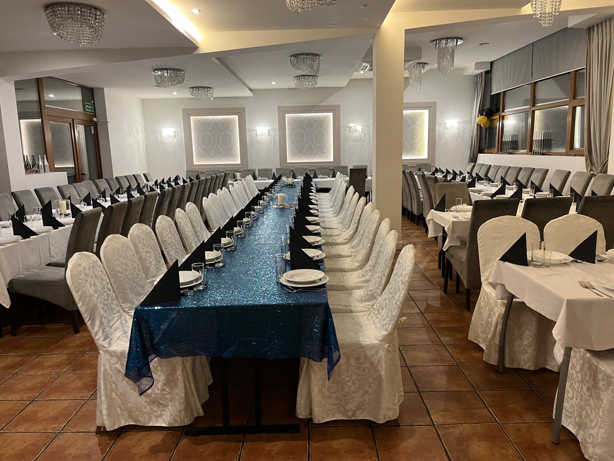 Uroczysta sala z długim stołem udekorowanym błyszczącym, niebieskim obrusem, nakrytym białymi talerzami i czarnymi serwetkami, krzesła w białych pokrowcach, kryształowe żyrandole