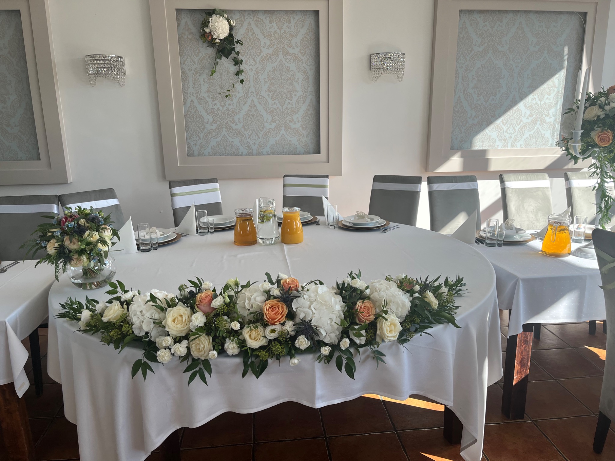 Uroczysta dekoracja stołu: biały obrus, kompozycja z białych i brzoskwiniowych róż, hortensji i zieleni, szklane naczynia i dzbanki z napojem, krzesła z szarymi pokrowcami, jasne wnętrze...