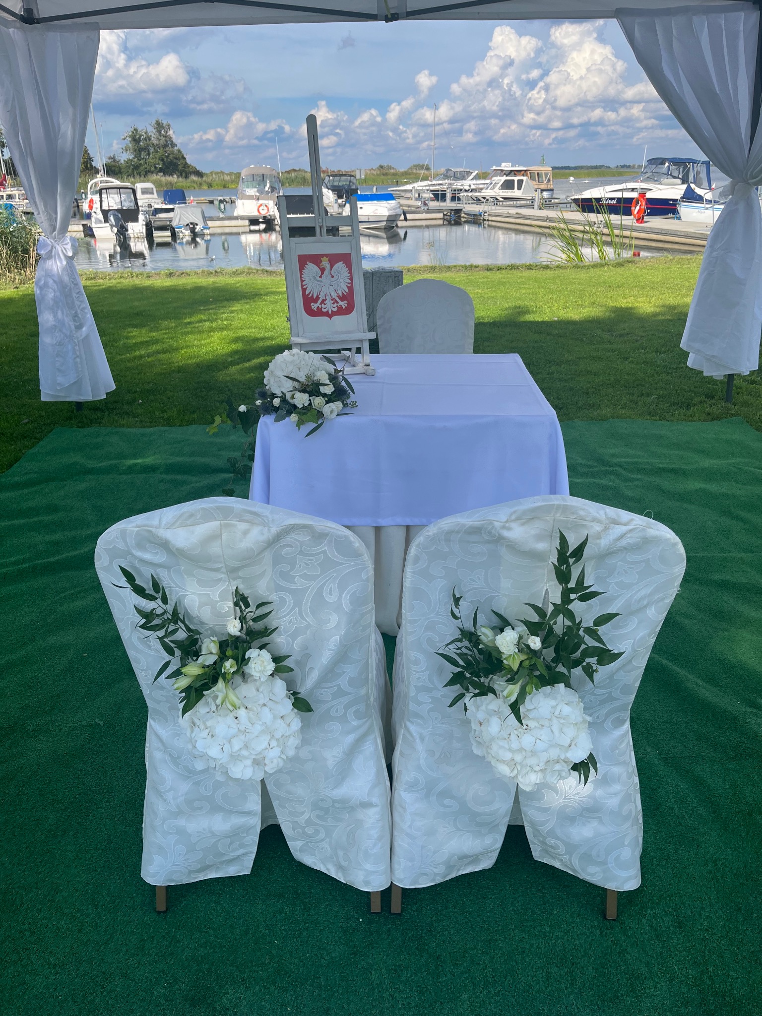 Dekoracja miejsca ceremonii ślubnej na zewnątrz: biały stół, krzesła w pokrowcach z kwiatami, godło Polski na sztaludze, widok na przystań jachtową w tle.