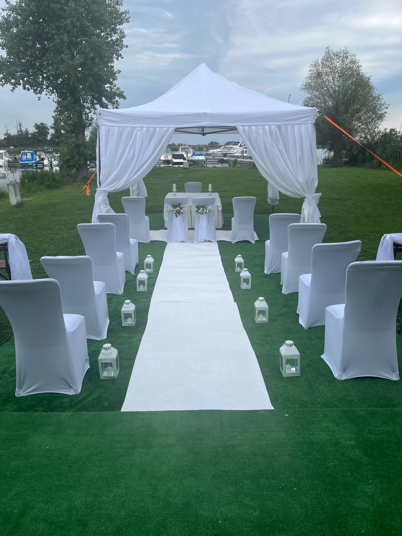 Dekoracja ślubna plenerowa: biały namiot z draperiami, krzesła w pokrowcach, biały dywan, lampiony, tło: jezioro i zieleń.