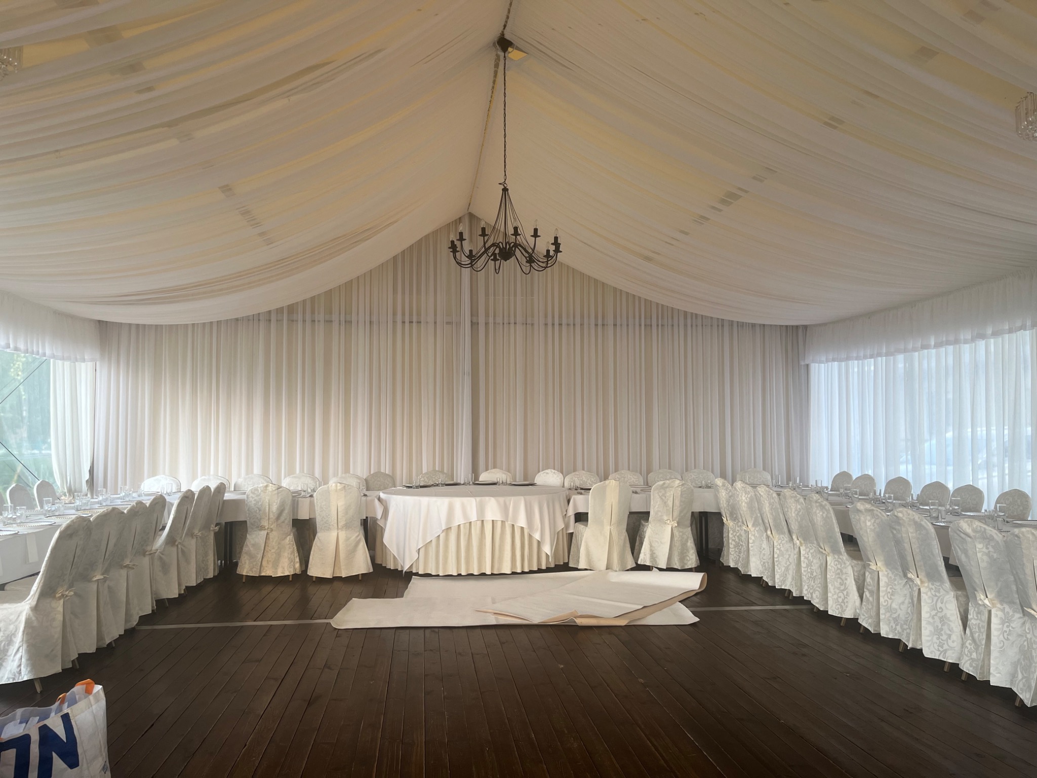 Sala bankietowa w trakcie przygotowań, białe krzesła w pokrowcach, nakryte stoły, dekoracja materiałowa pod sufitem, ciemny parkiet, rozłożone materiały na podłodze.
