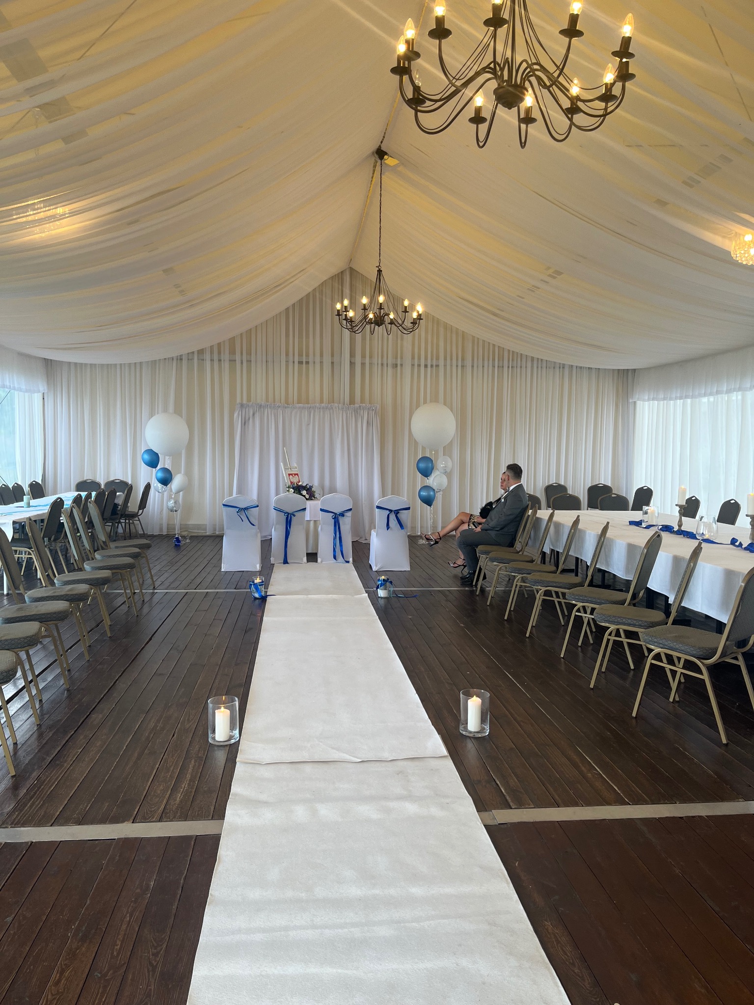 Uroczysta sala z białym dywanem, krzesłami udekorowanymi niebieskimi wstążkami, balonami i parą siedzącą przy stole, oświetlona żyrandolami.