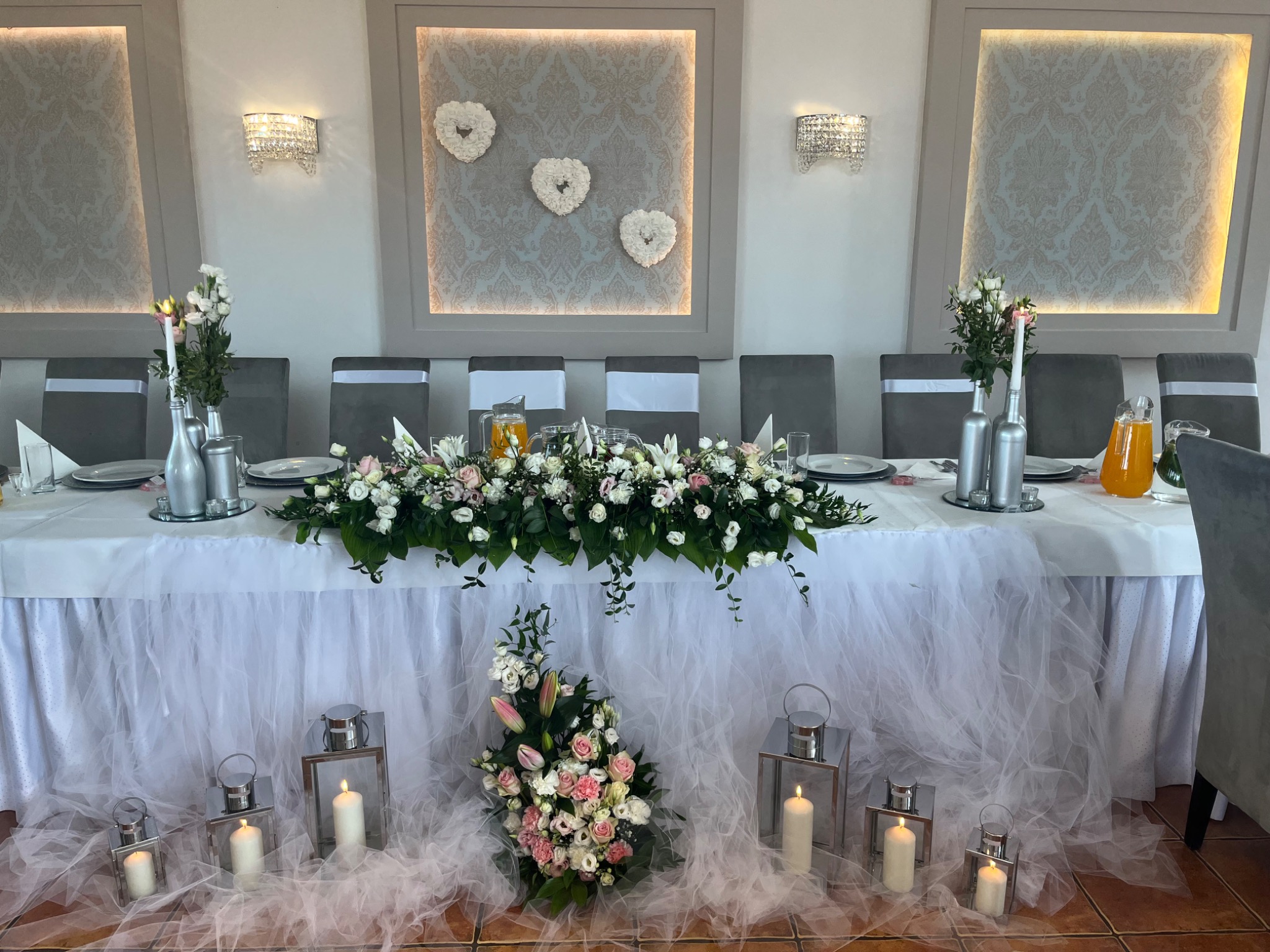 Dekoracja stołu weselnego z białymi kwiatami, srebrnymi lampionami i świecami, tło z ozdobnymi panelami i kryształowymi kinkietami.