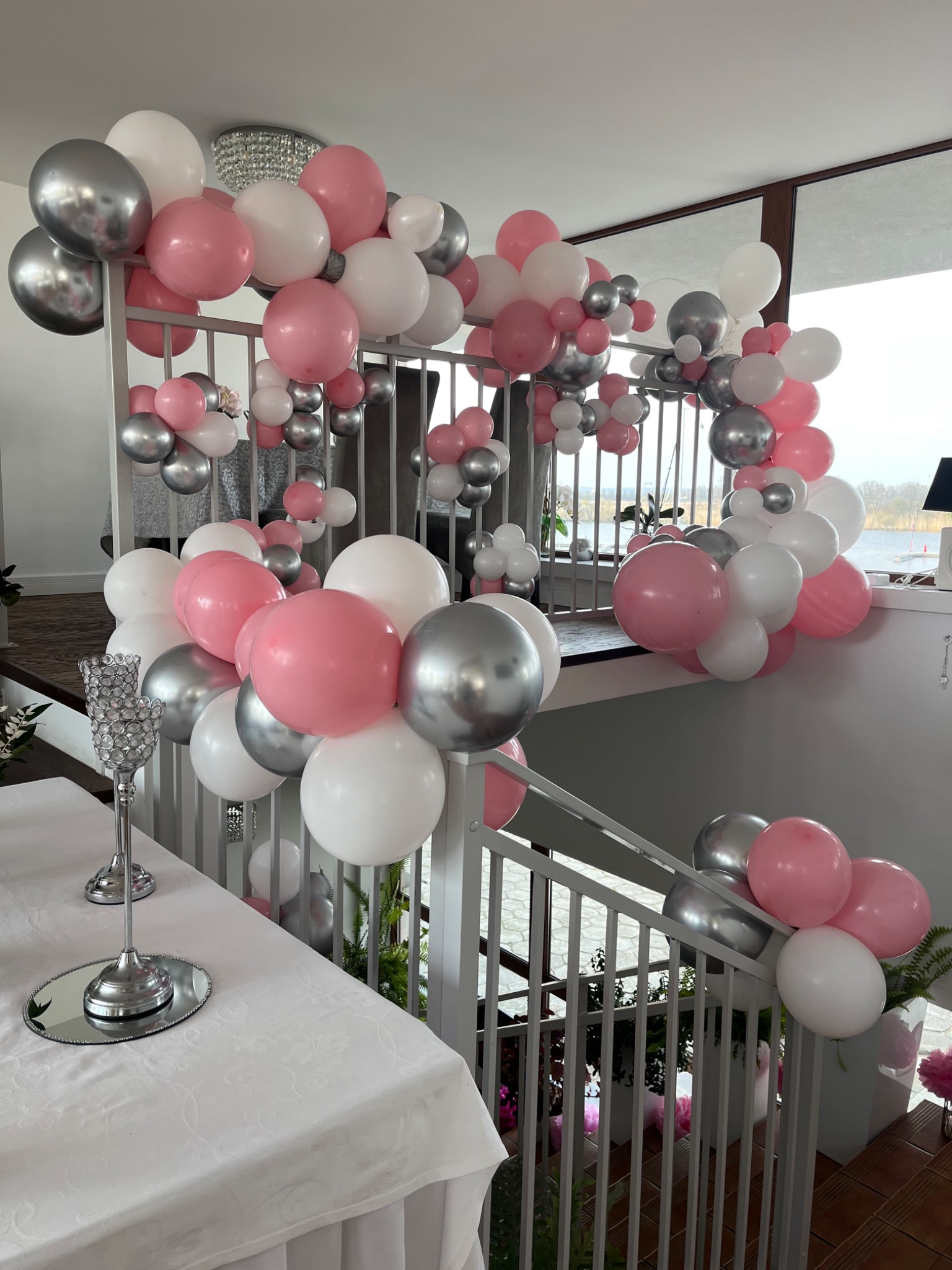 Dekoracja schodów na przyjęciu: girlanda z białych, różowych i srebrnych balonów, szklane świeczniki na stole z białym obrusem, widok z balkonu na wodę.