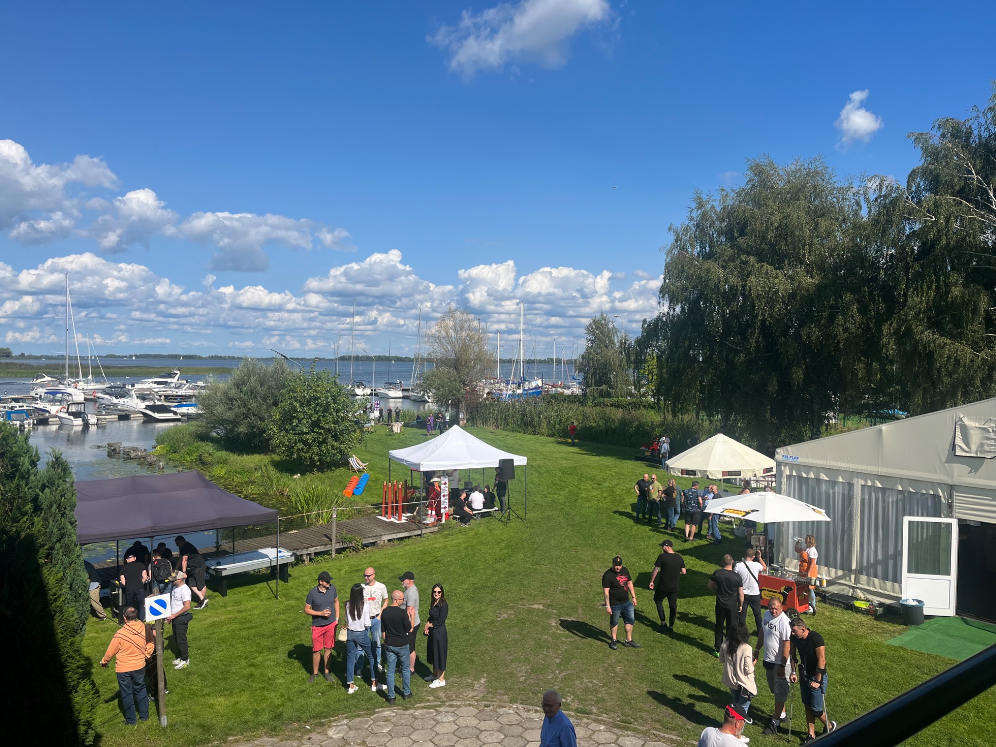 Plenerowa impreza firmowa nad jeziorem, z widocznymi namiotami, grupami ludzi rozmawiającymi i jachtami na wodzie w tle.