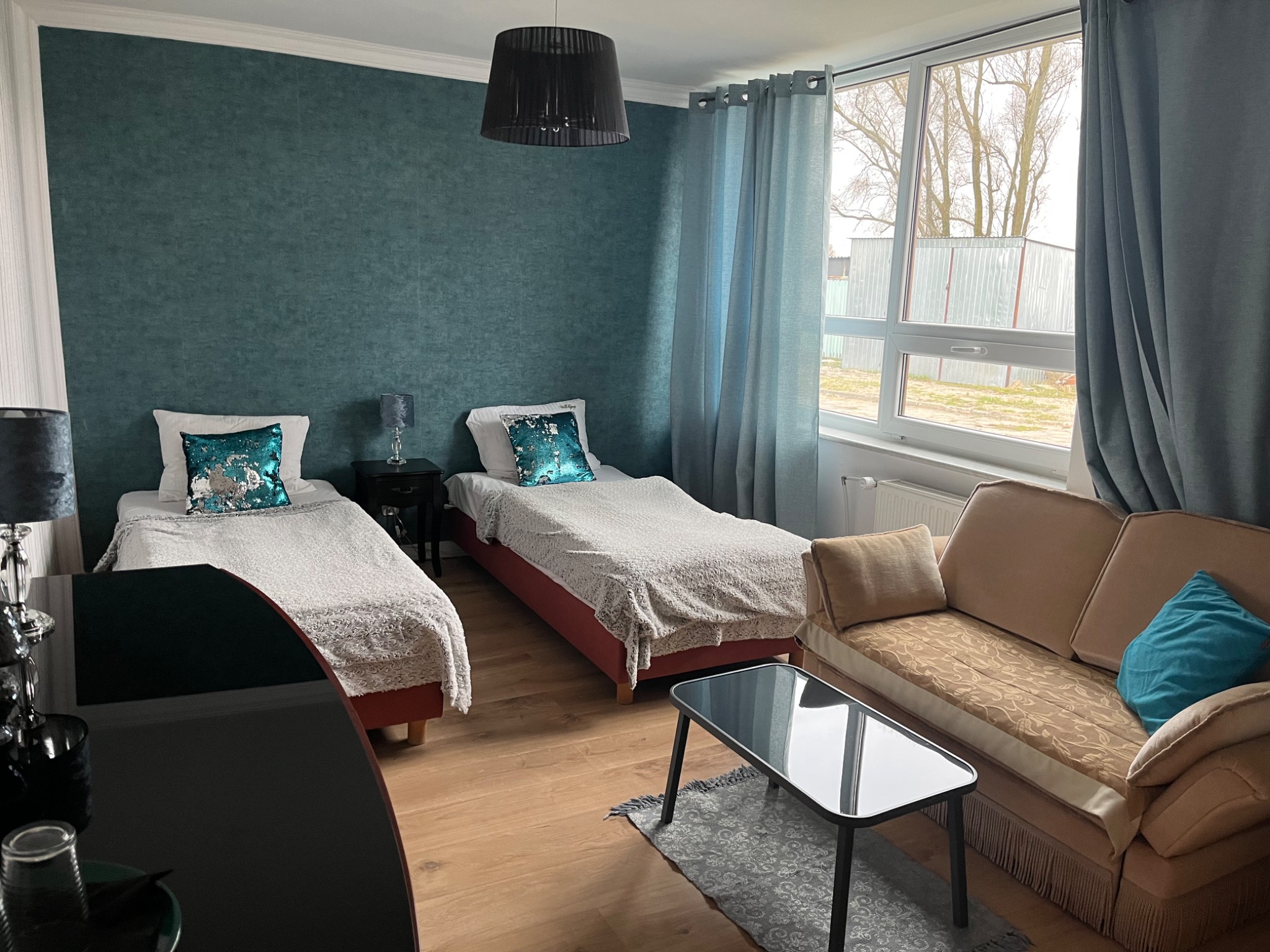 Pokój z dwoma łóżkami, turkusową ścianą i beżową sofą pod oknem, minimalistyczny stolik kawowy na dywaniku.