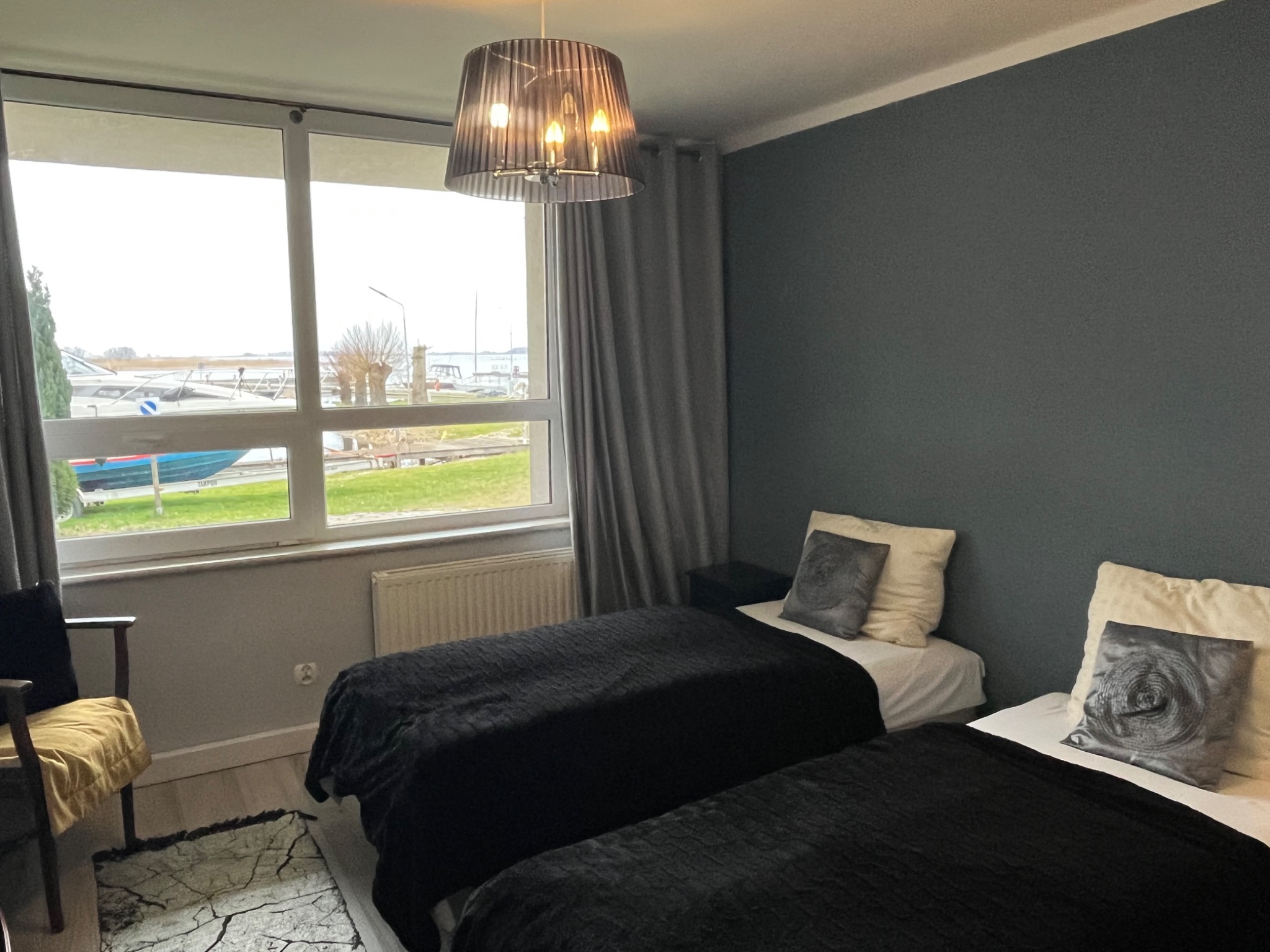 Pokój z dwoma łóżkami w Szczecinie, widok na przystań jachtową przez okno, ciemne ściany i czarne narzuty, lampa wisząca nad łóżkami.