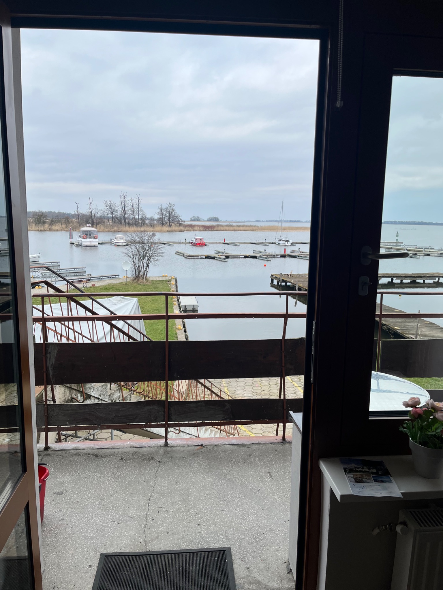 Widok z balkonu na przystań jachtową w pochmurny dzień. Na pierwszym planie fragment balkonu z betonową podłogą i metalową barierką. W tle woda, pomosty i kilka zacumowanych łodzi.