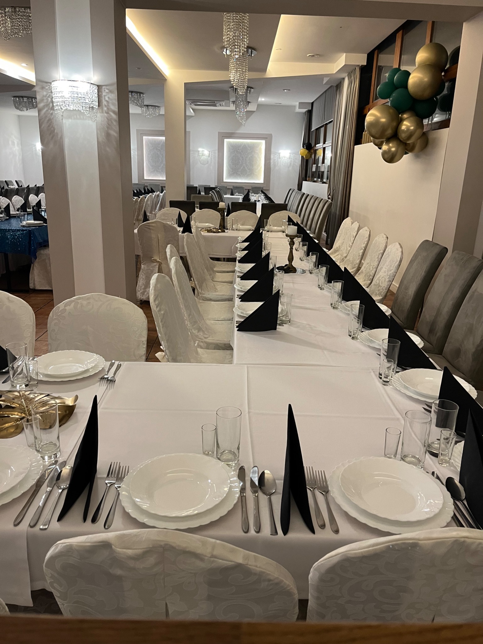 Uroczysta sala z długim stołem nakrytym białym obrusem, czarne serwetki, elegancka zastawa, krzesła z białymi pokrowcami i szare krzesła, dekoracja z zielono-złotych balonów, kryształowe żyrandole.