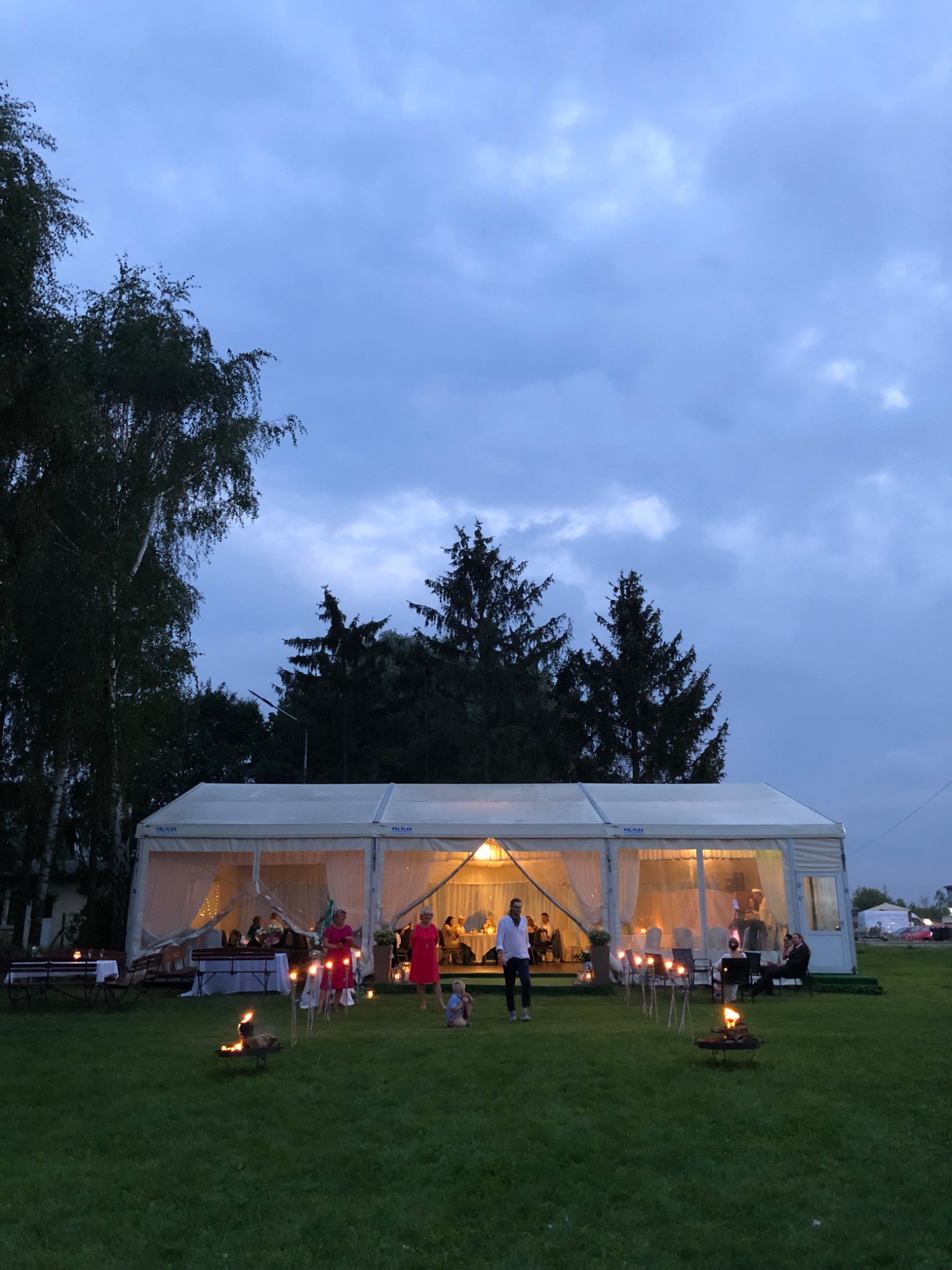 Uroczystość plenerowa pod białym namiotem z oświetleniem, goście wewnątrz, płonące pochodnie na zewnątrz, wieczorne niebo w tle.