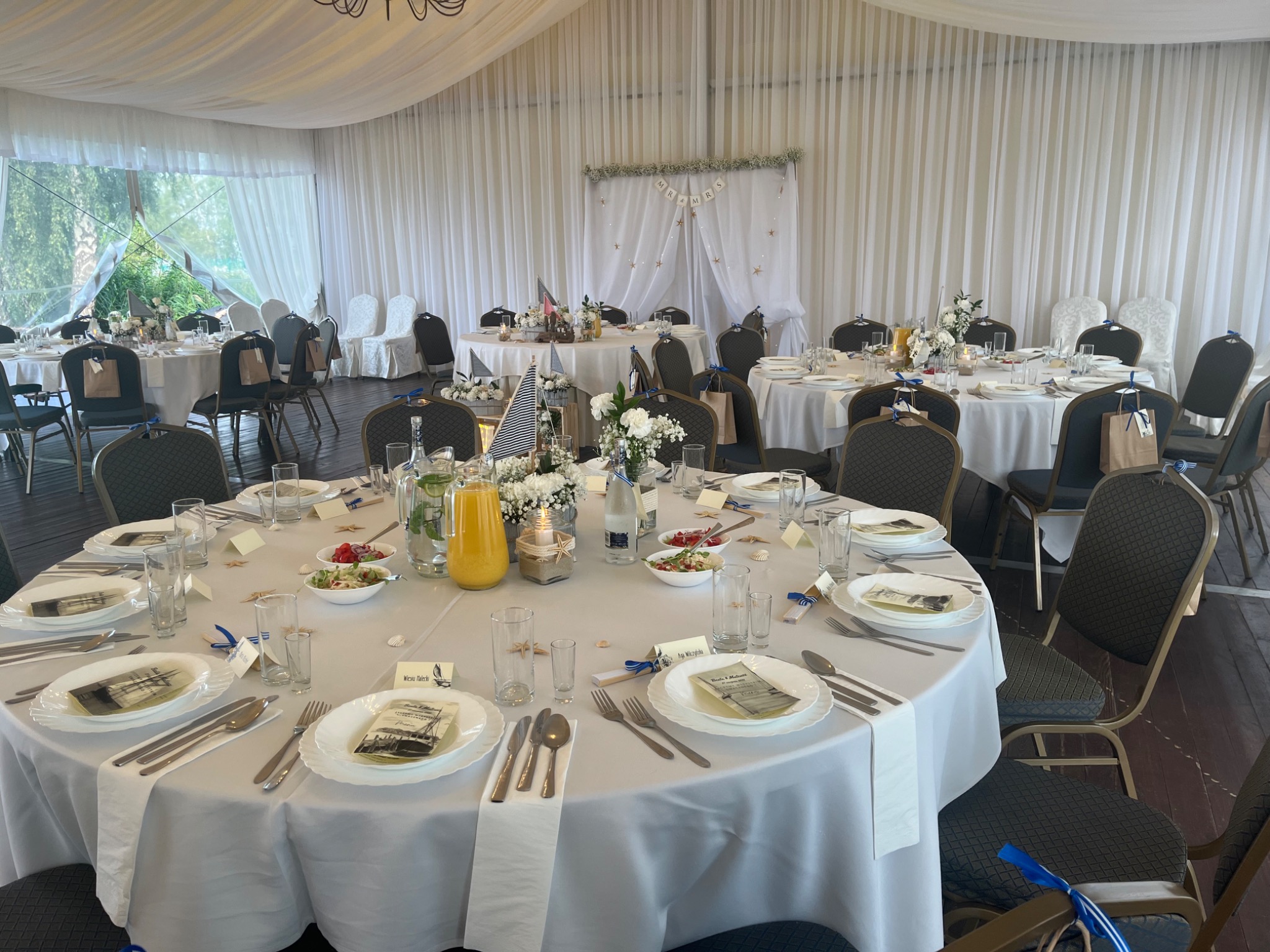 Uroczysta sala bankietowa z okrągłymi stołami nakrytymi białymi obrusami, dekoracjami w stylu marynistycznym i krzesłami z niebieskimi wstążkami, przygotowana na przyjęcie weselne lub inną...