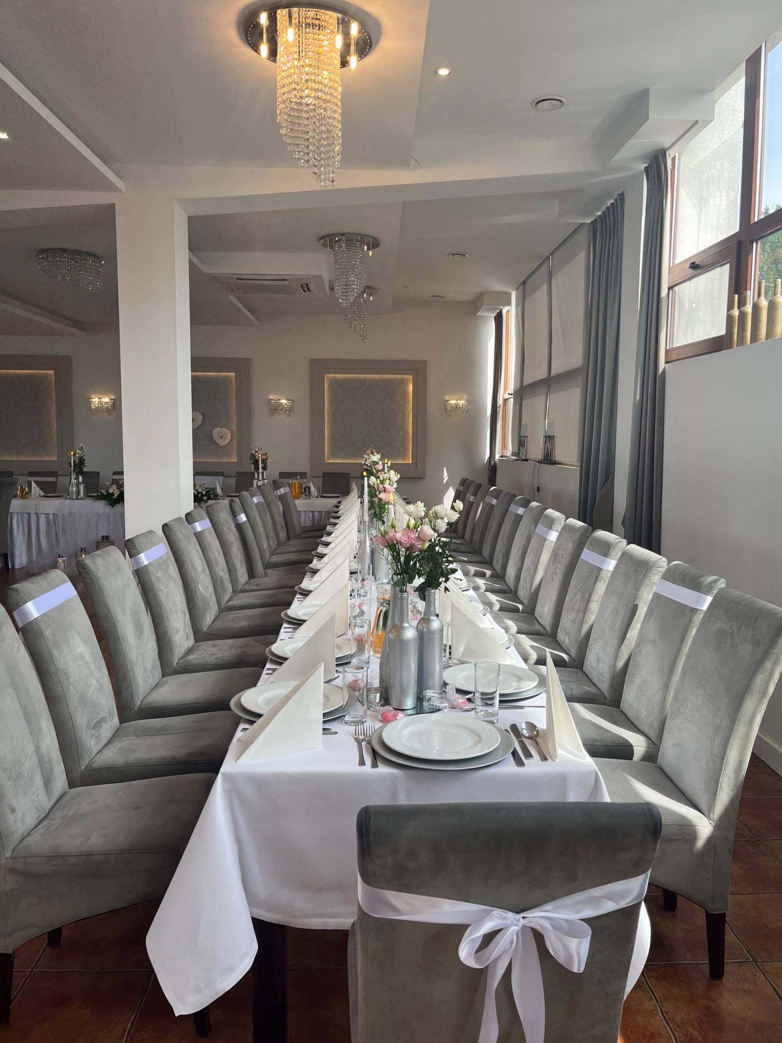 Długi, elegancko nakryty stół weselny z szarymi krzesłami ozdobionymi białymi wstążkami, srebrnymi wazonami z kwiatami i kryształowymi żyrandolami w jasnym wnętrzu sali bankietowej.