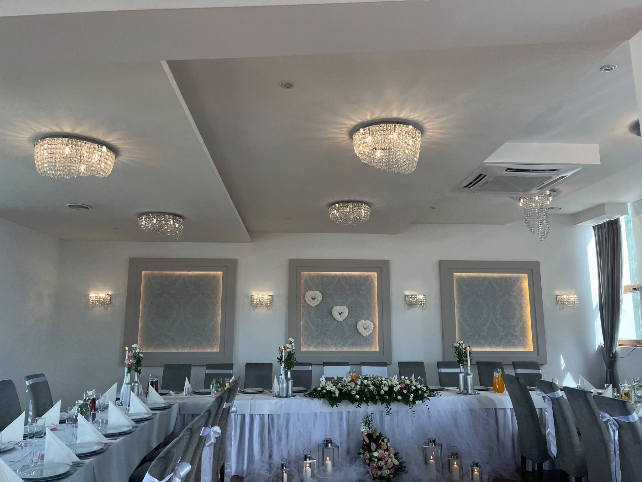 Elegancko udekorowany stół weselny z kwiatami, świecami w lampionach i szarymi krzesłami, oświetlony kryształowymi żyrandolami, z dekoracyjnymi panelami ściennymi w tle.