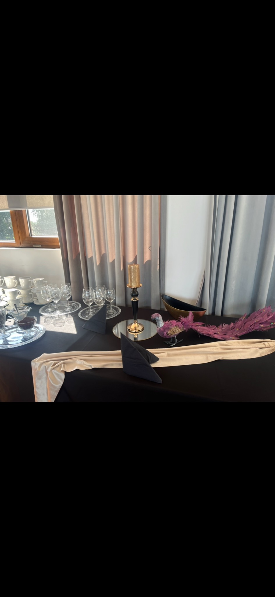 Elegancko nakryty stół z czarnym obrusem, złotym bieżnikiem, zastawą stołową, świecznikiem i dekoracyjnym pawiem z piórami.