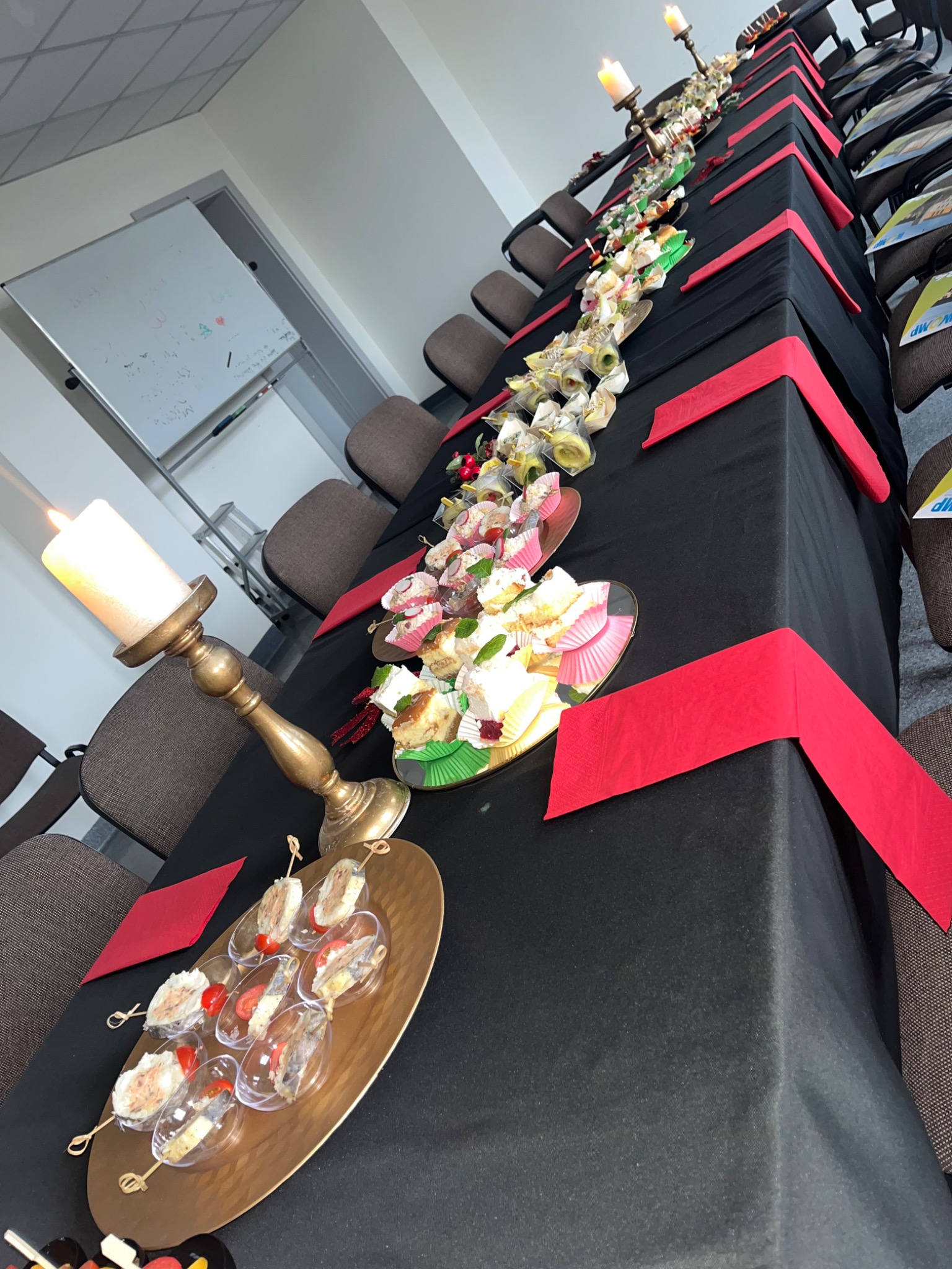 Długi stół nakryty czarnym obrusem z czerwonymi dekoracjami, zastawiony przekąskami, ciastami i świecami w złotych świecznikach, przygotowany na spotkanie firmowe.