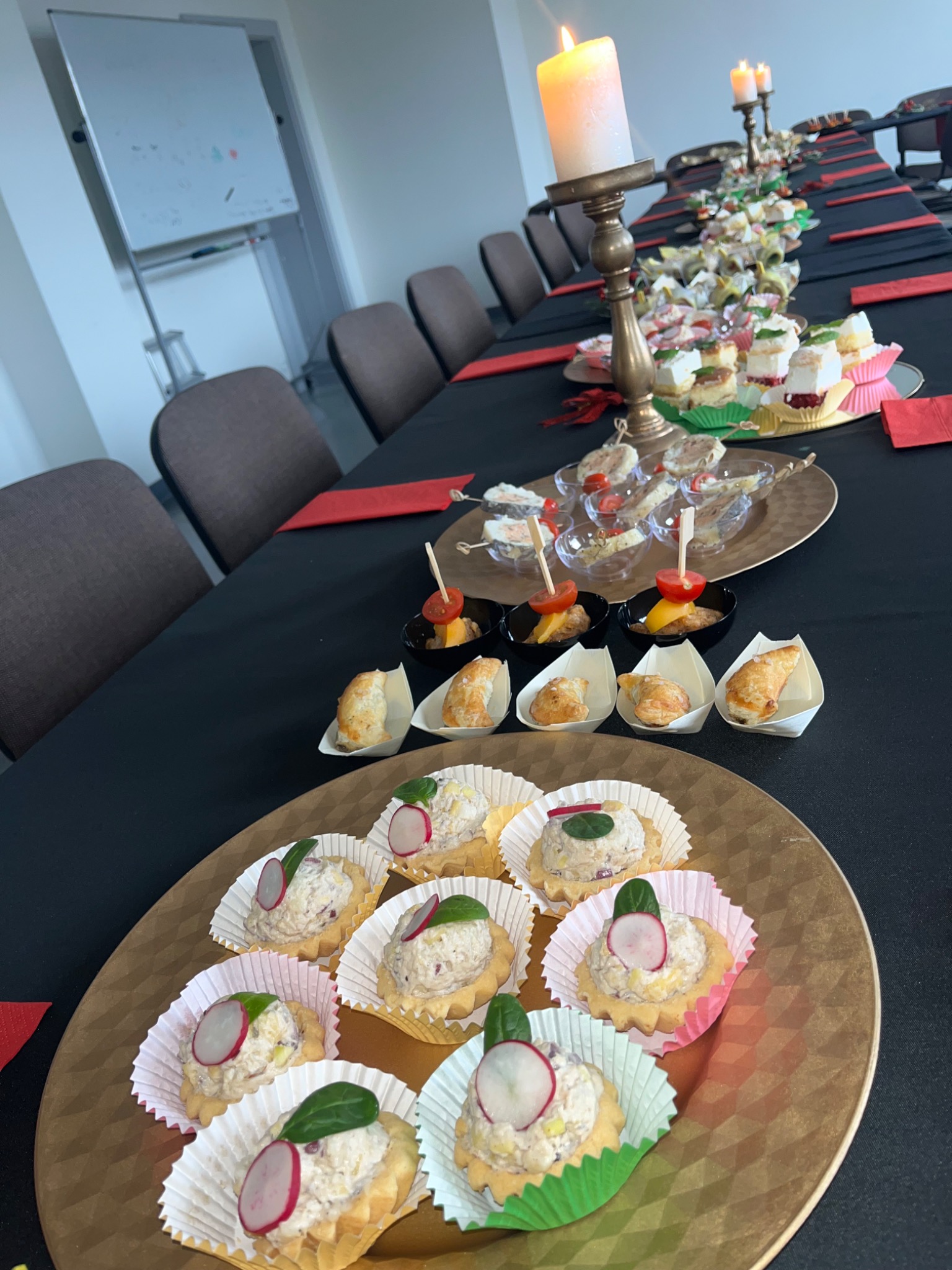 Uroczysty stół konferencyjny z czarnym obrusem, ozdobiony złotymi świecznikami ze świecami i różnorodnymi przekąskami na złotych tacach, w tym tartaletki z pastą i rzodkiewką, mini-szaszłyki...