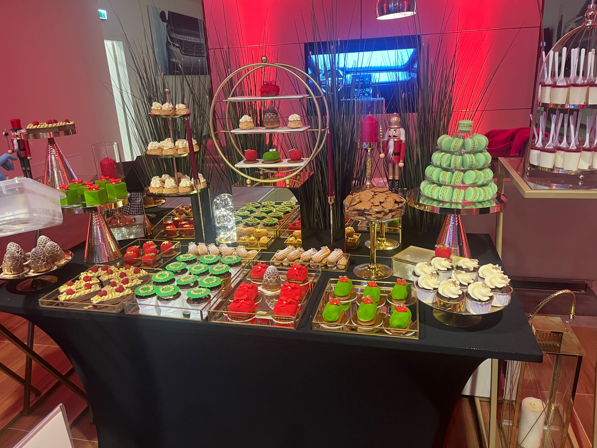 Bogato zastawiony stół z różnorodnymi deserami: babeczki z kremem, makaroniki, ciastka w kształcie choinek i inne słodkości, dekoracje świąteczne, czerwone oświetlenie w tle.