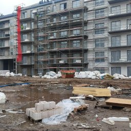 DACHTECHNIK PREMIUM SPÓŁKA Z OGRANICZONĄ ODPOWIEDZIALNOŚCIĄ - Budynek w trakcie ocieplania elewacji, rusztowania na całej wysokości, widoczne materiały izolacyjne i pracownicy. Elementy w kolorze czerwonym na elewacji.