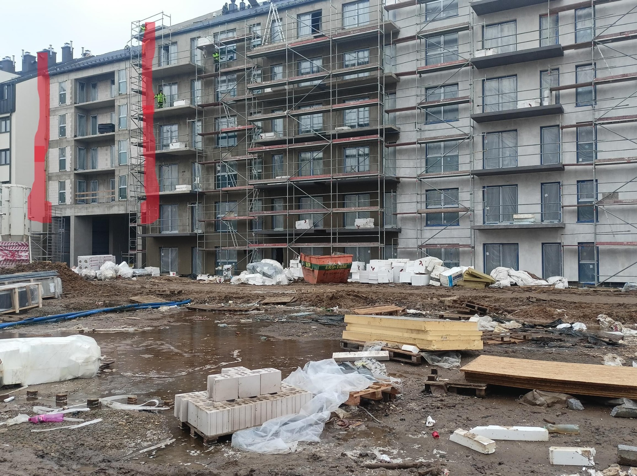 Budynek w trakcie ocieplania elewacji, rusztowania na całej wysokości, widoczne materiały izolacyjne i pracownicy. Elementy w kolorze czerwonym na elewacji.