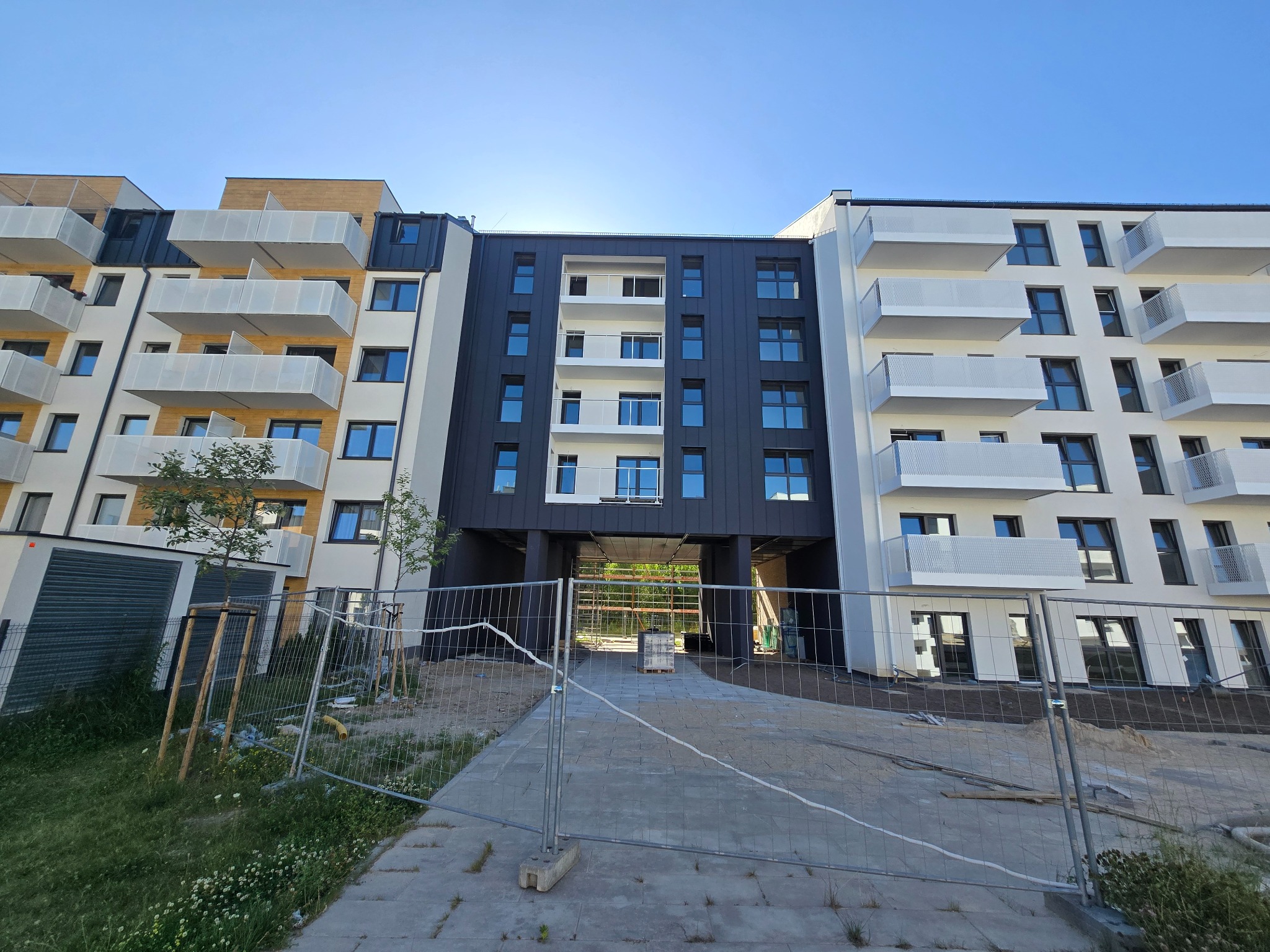 Nowoczesny budynek wielorodzinny z balkonami, wykończony w bieli, czerni i drewnie, widoczny przez ogrodzenie budowlane. Ujęcie z dołu, błękitne niebo.