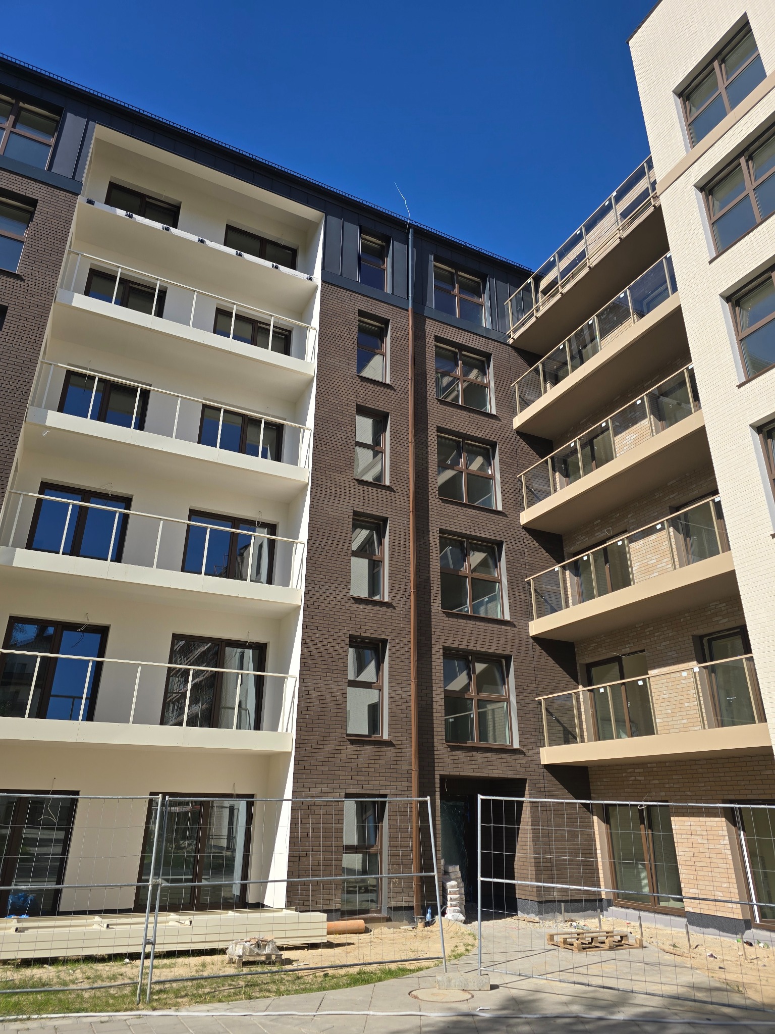 Nowoczesna elewacja budynku wielorodzinnego z balkonami, wykończona cegłą klinkierową w różnych odcieniach brązu i beżu, pod błękitnym niebem. Częściowe ogrodzenie terenu.