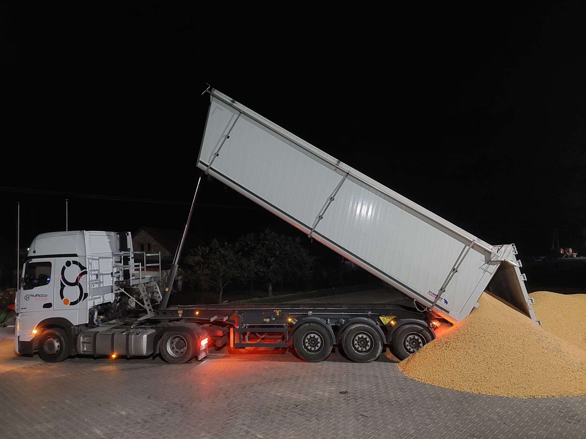 Biała ciężarówka z naczepą wywrotką, przechylona w trakcie rozładunku jasnych ziaren na ciemnym tle nocnego krajobrazu.