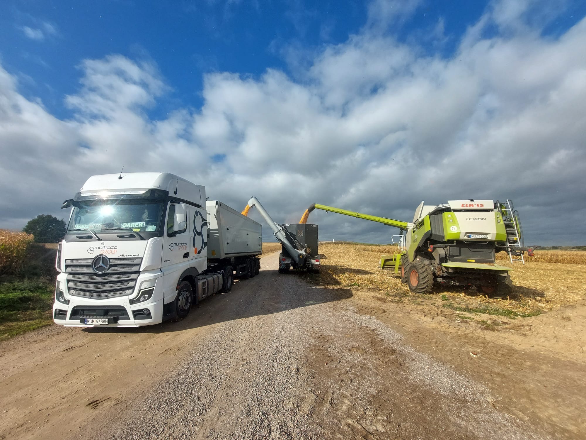 Kombajn zbożowy Claas Lexion przekazuje zebrane zboże do naczepy ciężarówki Mercedes-Benz Actros z logo Multicco Group na tle pochmurnego nieba.