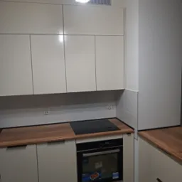 Kremowa zabudowa kuchenna z drewnianym blatem, płyta indukcyjna i piekarnik w zabudowie, widoczne gniazdka elektryczne na ścianie.