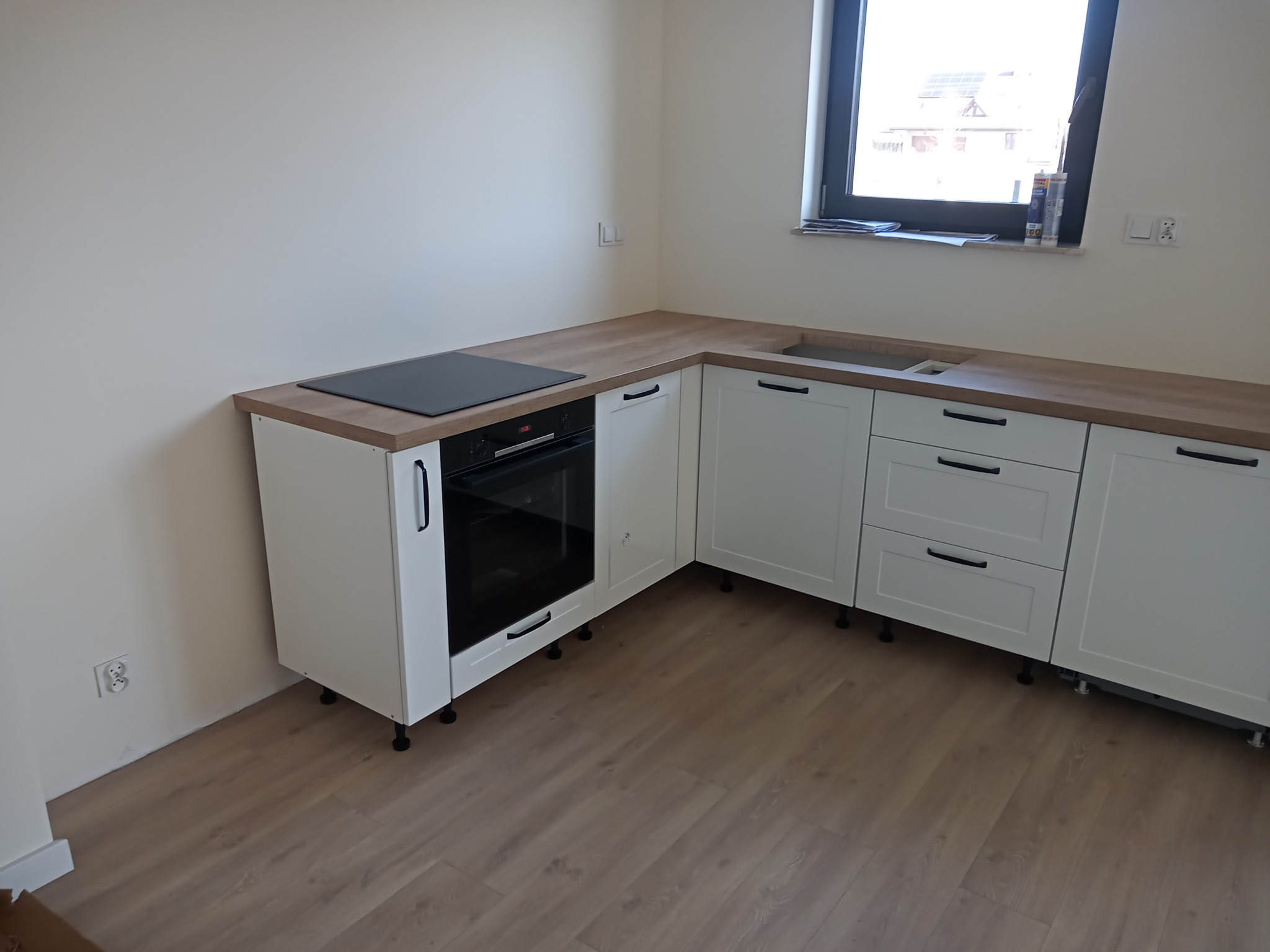 Jasna kuchnia z białymi szafkami i drewnianym blatem, widoczna płyta grzewcza i piekarnik, okno z widokiem na zabudowania na zewnątrz, wnętrze w stanie surowym przed wykończeniem.