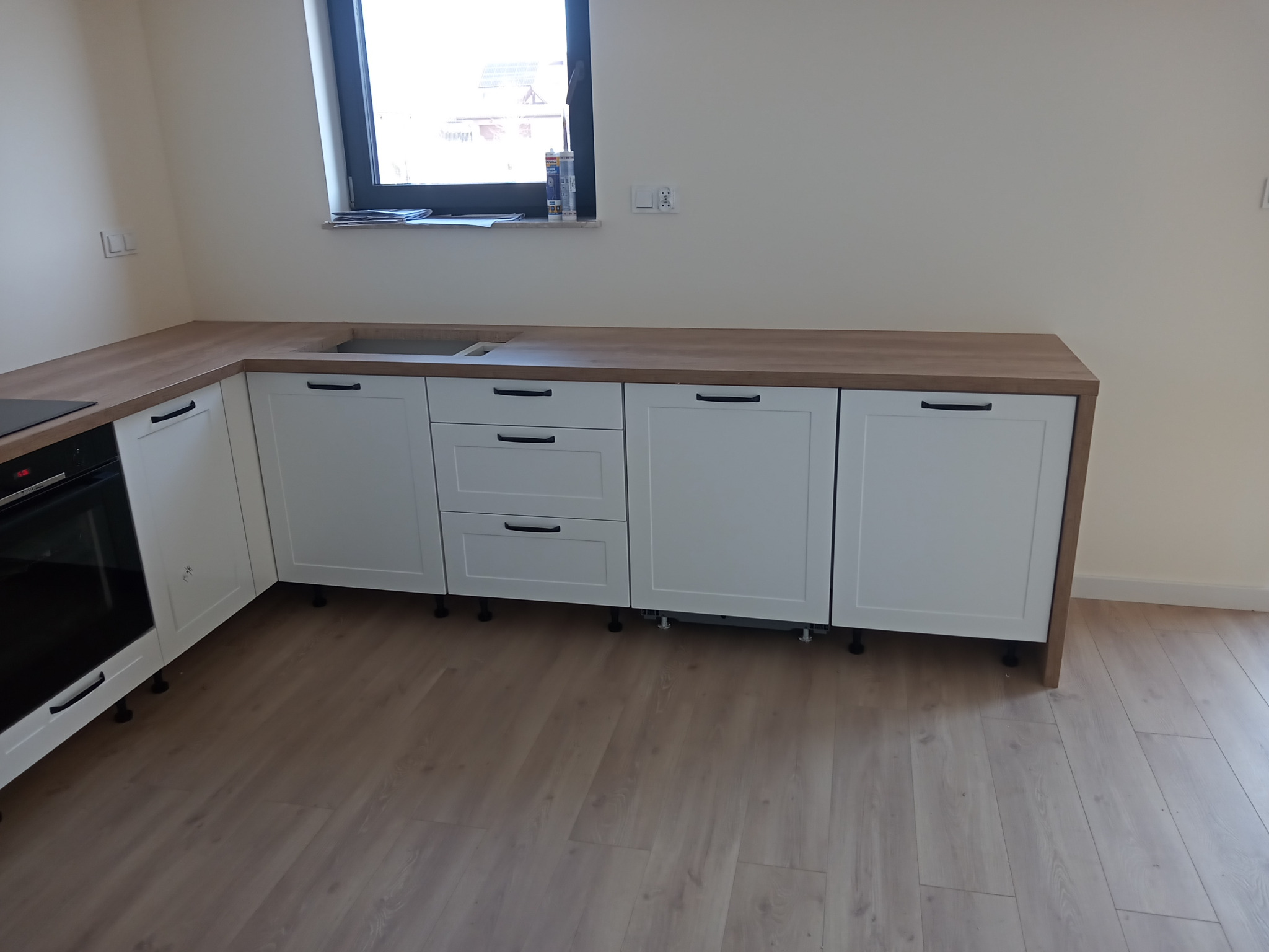 Nowoczesna, jasna kuchnia z białymi szafkami i drewnianym blatem, jeszcze bez sprzętów AGD, widoczna wnęka na zlew, silikon na parapecie okna.